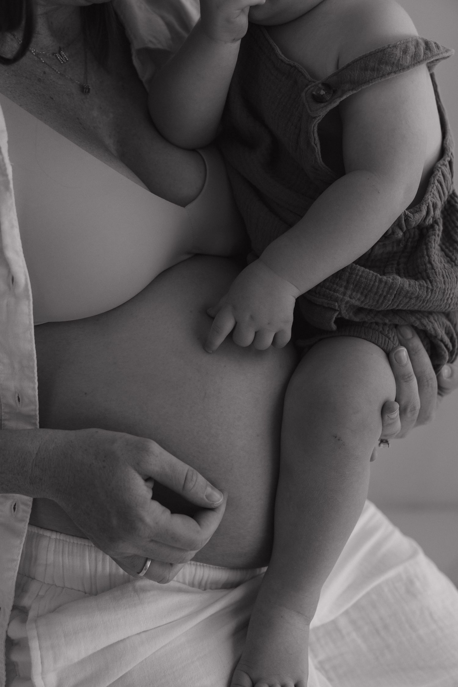 Black and white photo of pregnant woman holding baby on her bare bump, with partner's hand gently touching her side.