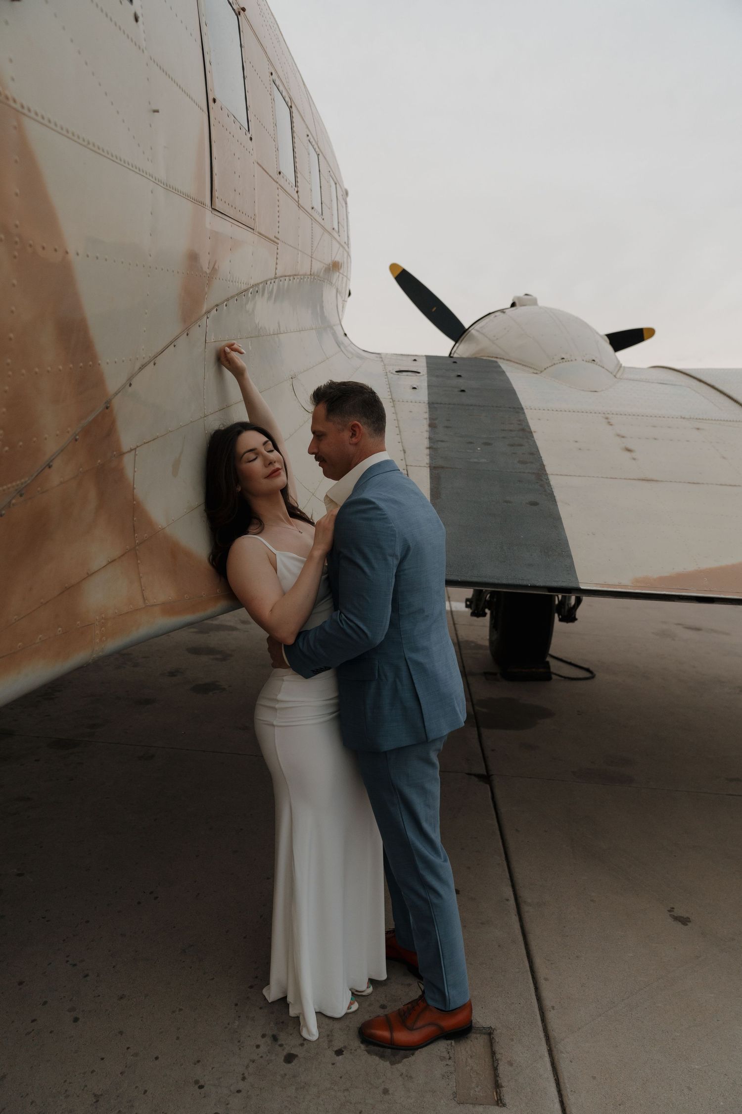 Couple embracing beside a vintage propeller aircraft during their engagement shoot at an airfield.