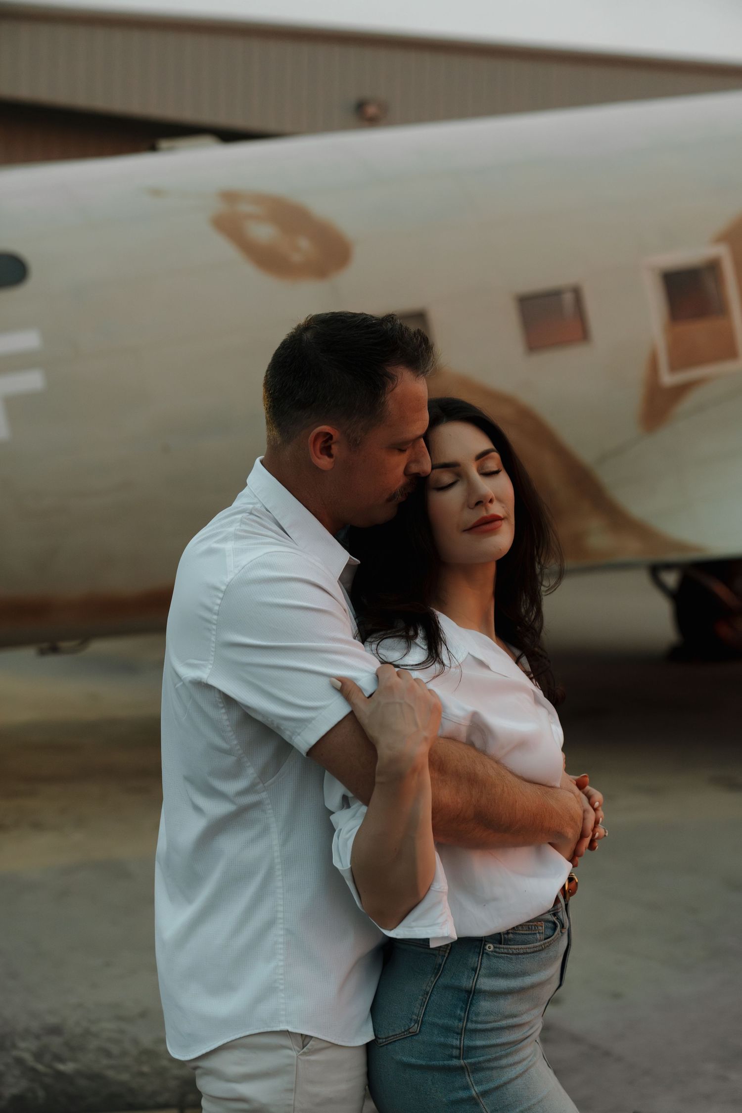 A couple embraces tenderly in front of a vintage airplane on a tarmac, both wearing white shirts.