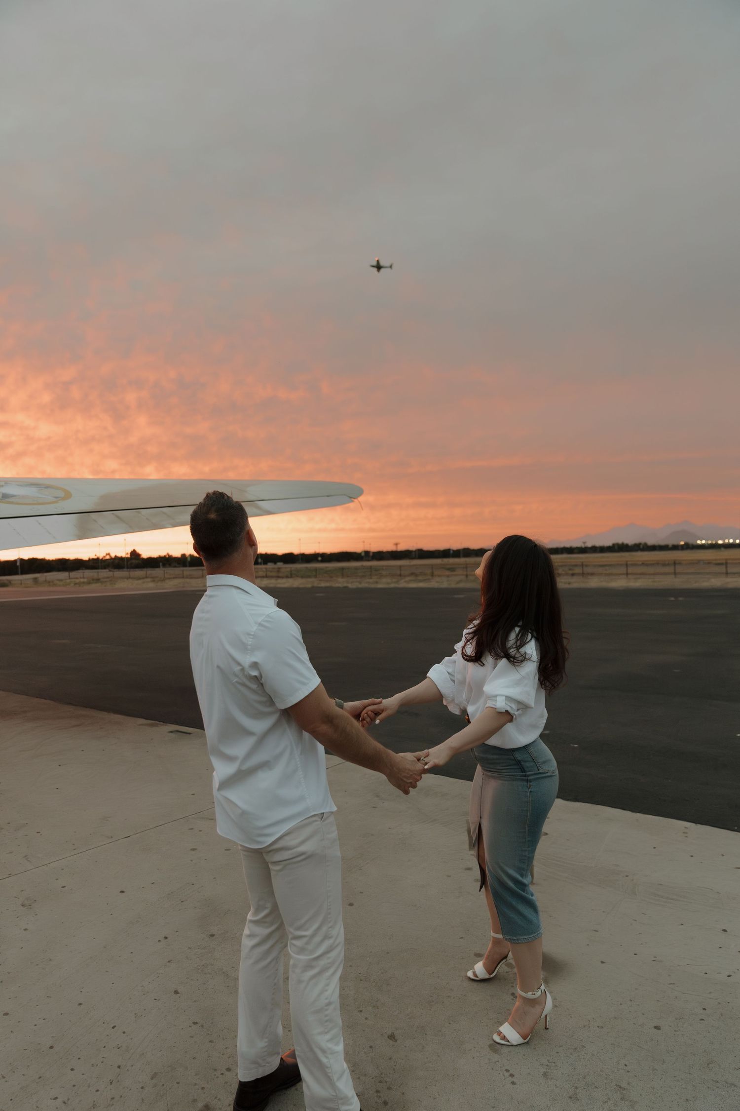 Couple holding hands on airport tarmac at sunset with airplane flying overhead and vibrant pink sky.