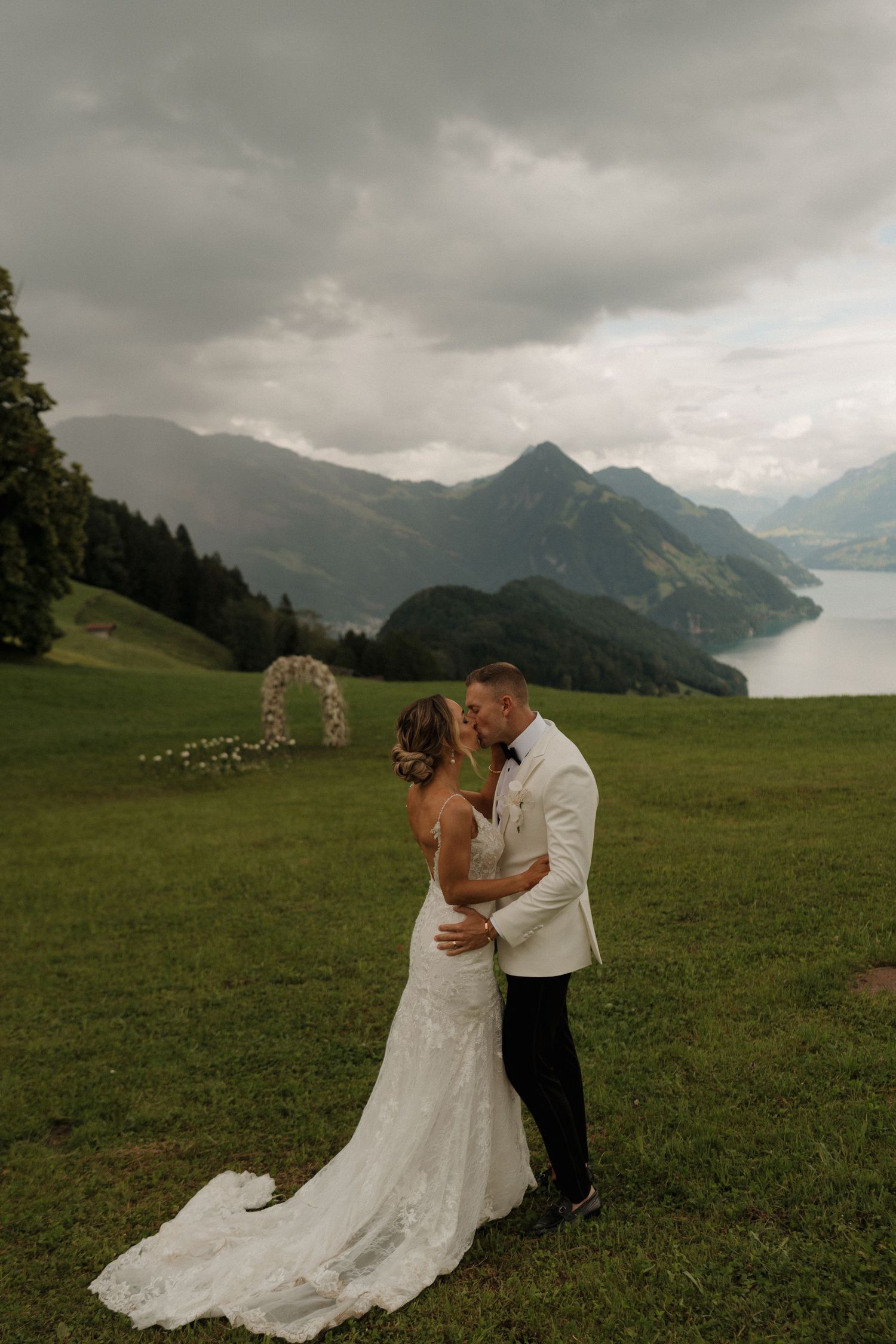 Newlyweds share a romantic kiss on green hillside with floral arch and stormy Swiss mountain lake backdrop.