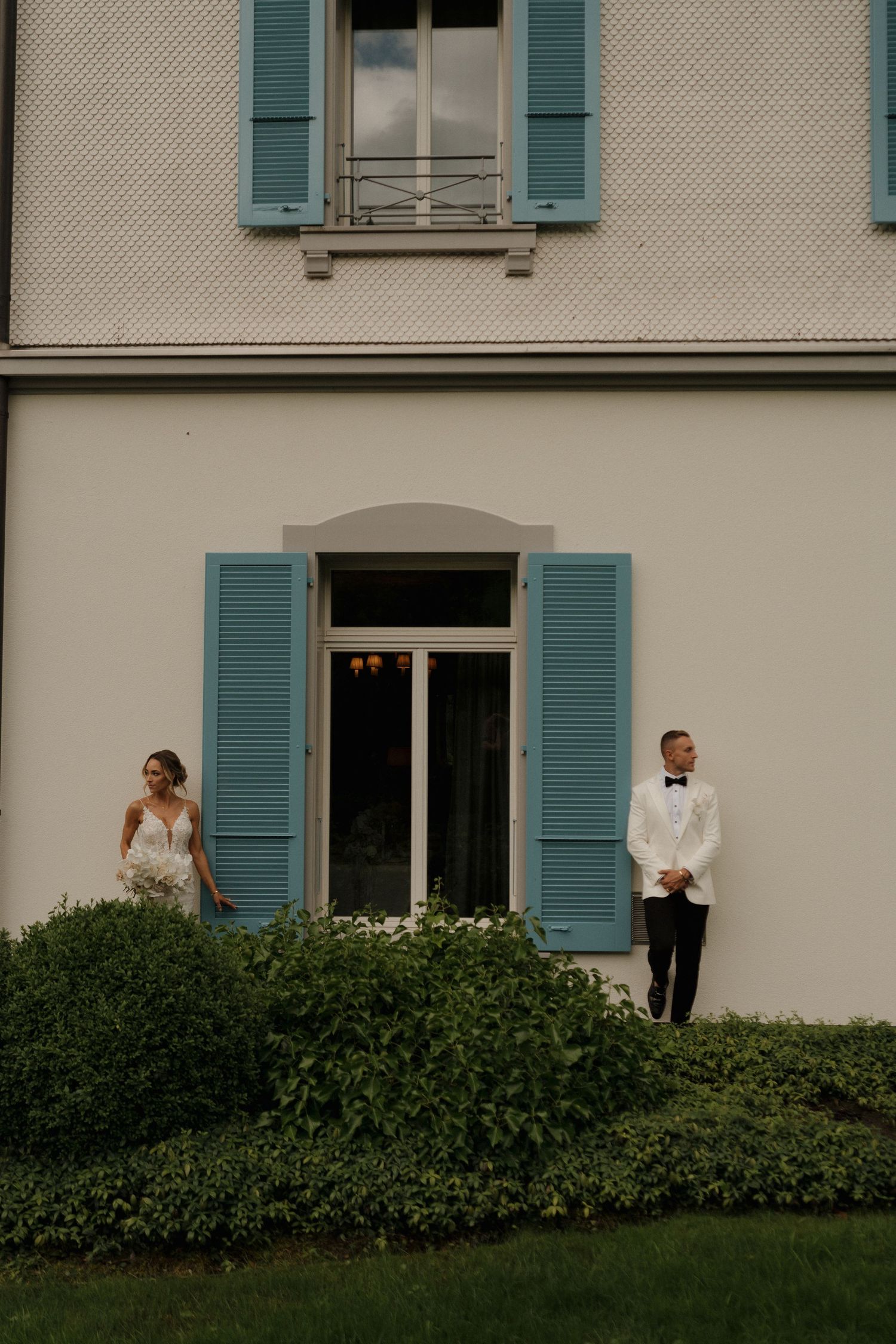 Bride and groom standing apart outside elegant villa with blue shutters on their wedding day.