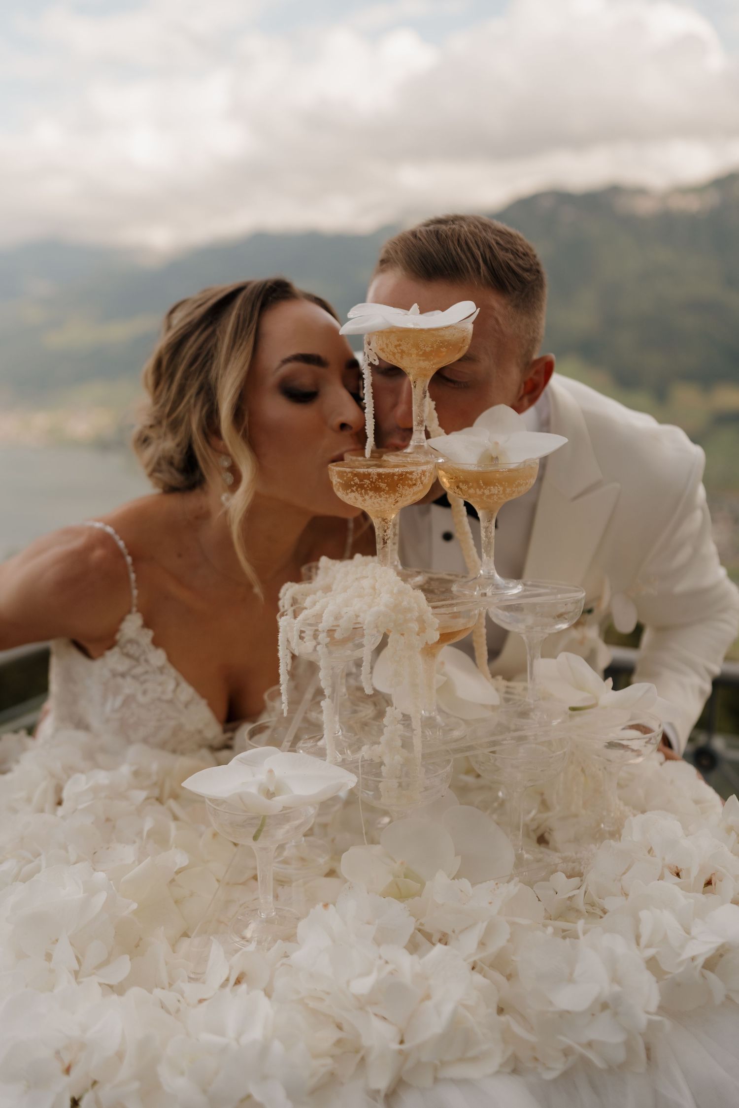 Bride and groom sipping champagne together from a tower of glasses with alpine mountains behind them.