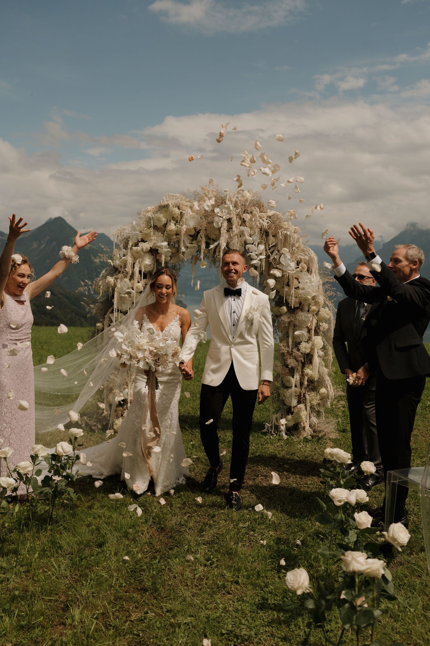 Joyful newlyweds exit outdoor mountain wedding ceremony as guests toss white flower petals in celebration.