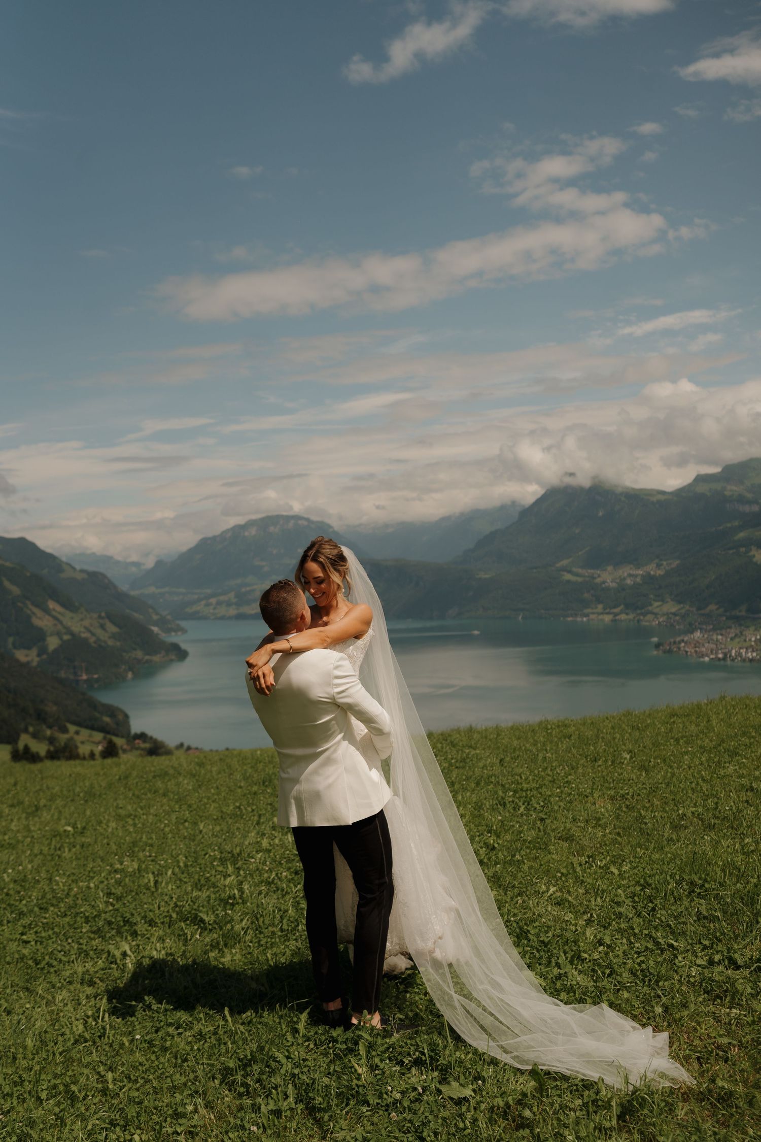 Bride and groom embrace on a green hillside with a stunning alpine lake and mountain backdrop on their wedding day.