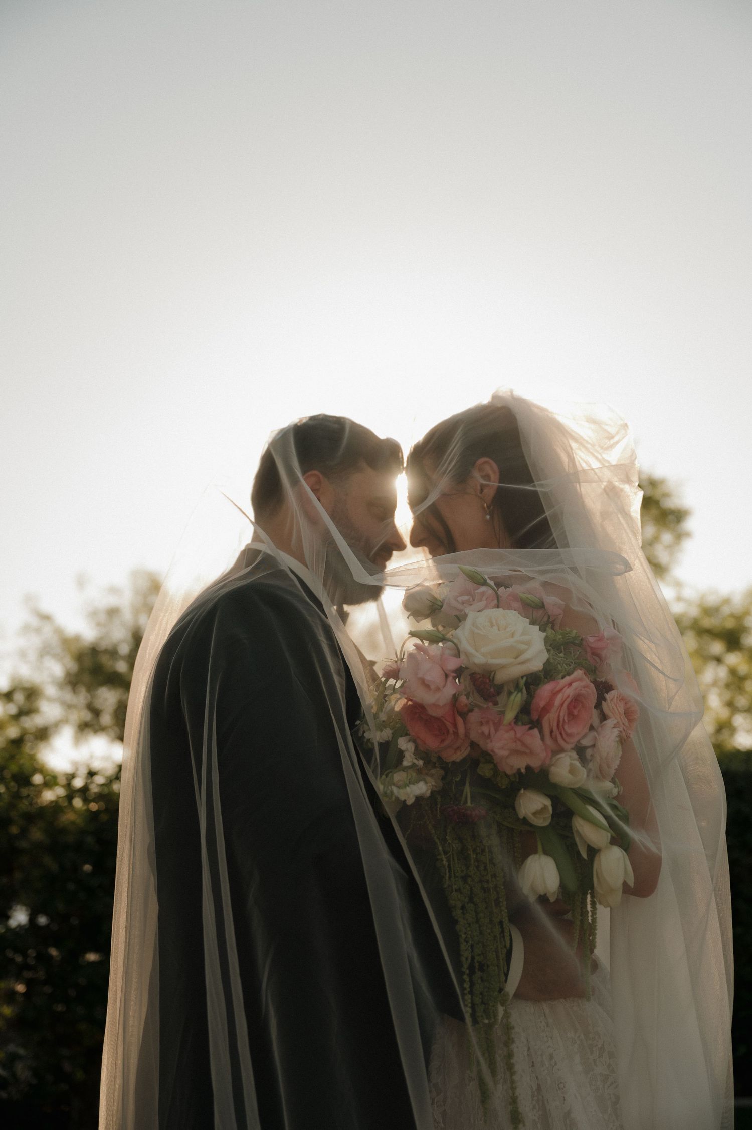 Romantic backlit photo of bride and groom sharing a kiss under flowing veil at golden hour sunset.