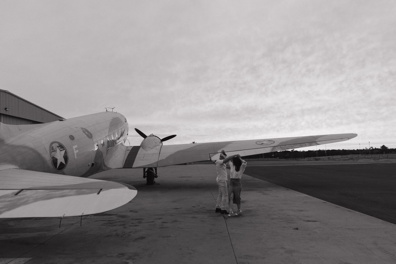 Black and white photo of a vintage WWII military aircraft on a tarmac with two people standing nearby.