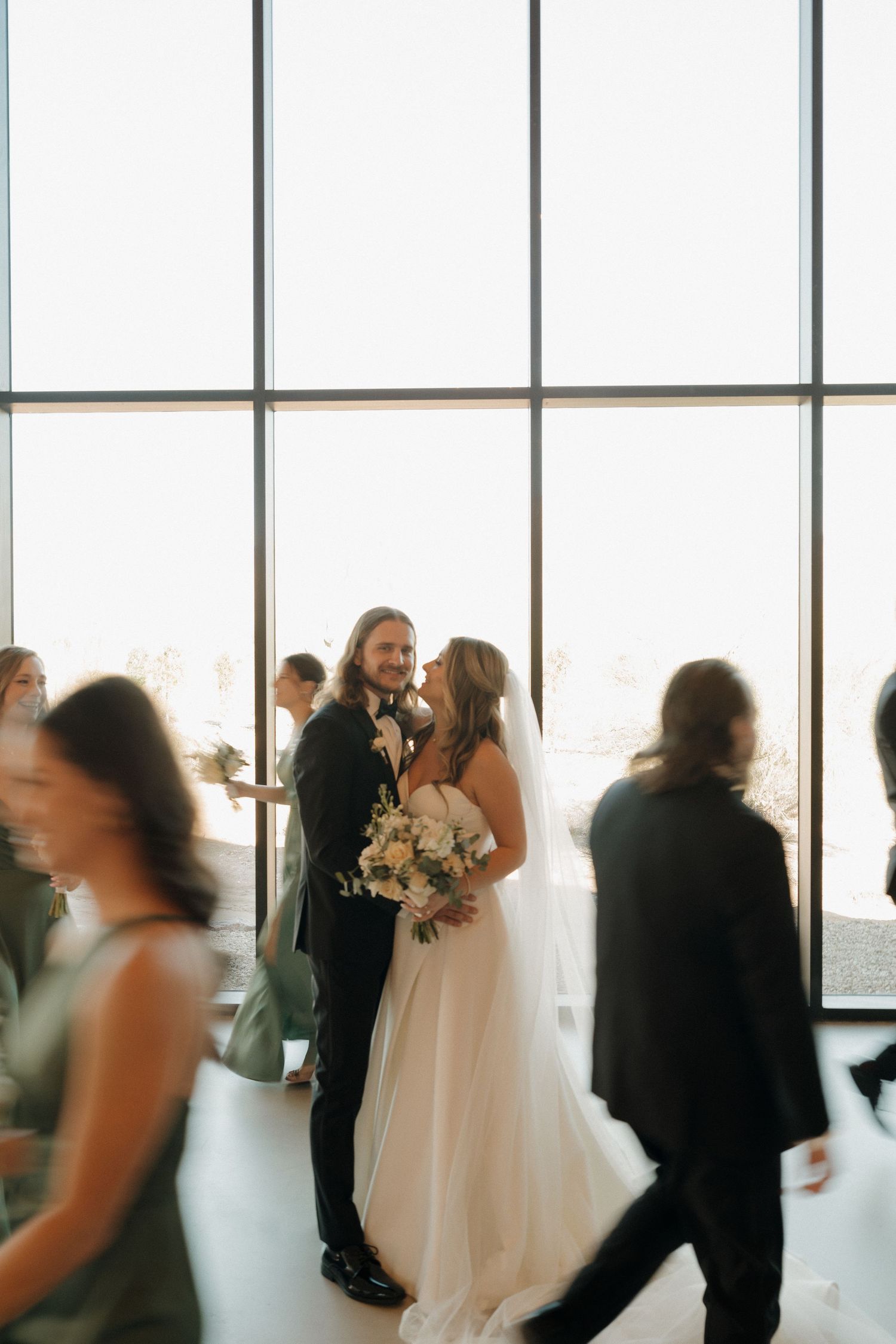 Bride and groom sharing romantic moment surrounded by guests in modern venue with floor windows.