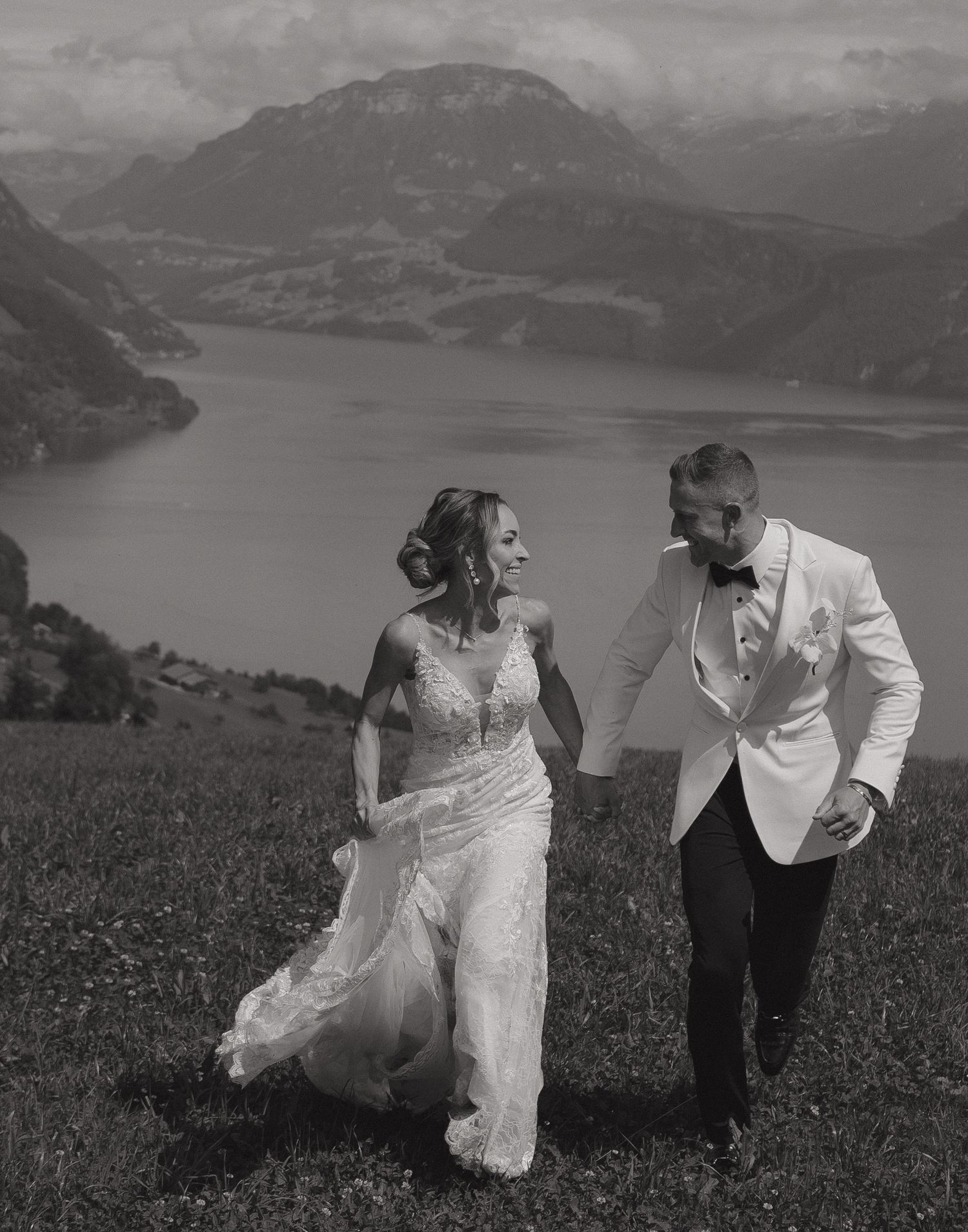 Black and white photo of smiling couple running through alpine meadow with dramatic lake and mountain scenery.