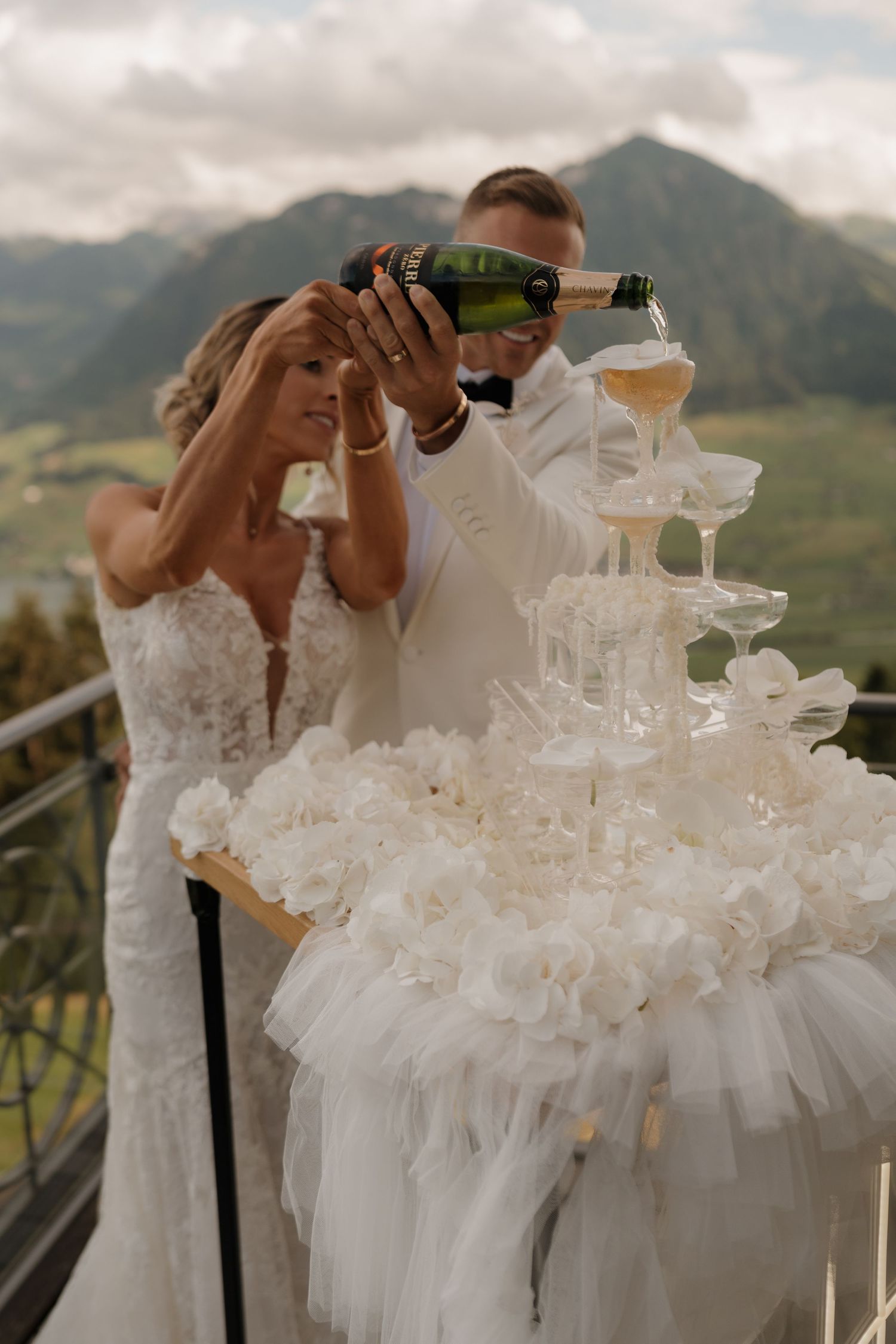 Bride and groom pouring champagne tower on a balcony with breathtaking mountain and lake scenery.