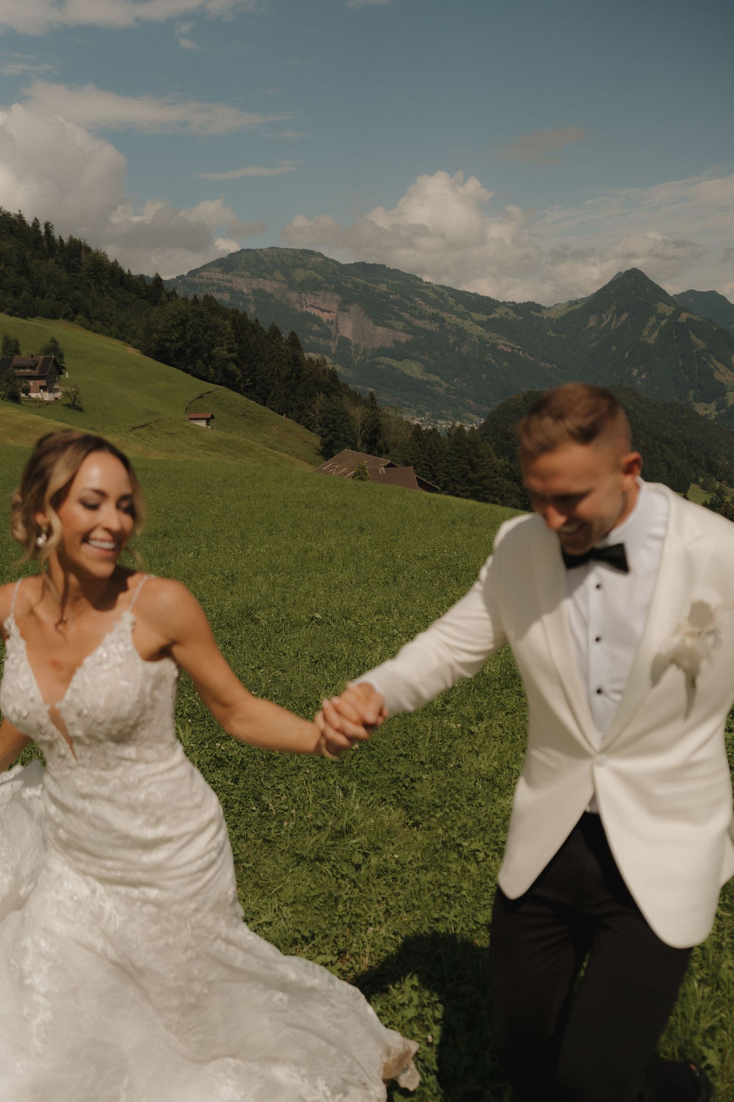 Joyful bride and groom holding hands running through green alpine meadow on their wedding day.