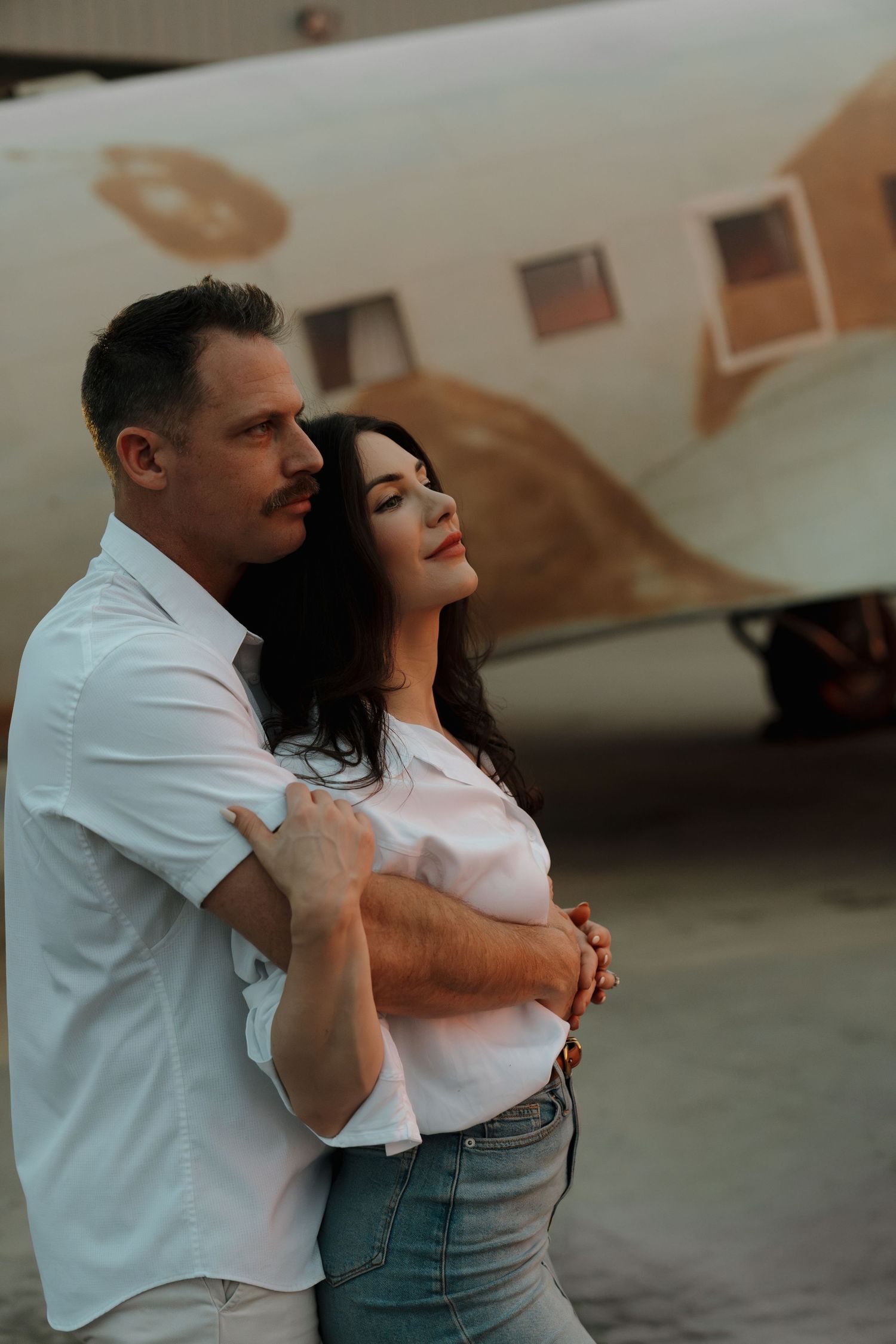 A couple in white shirts embraces romantically in front of a vintage airplane outdoors.
