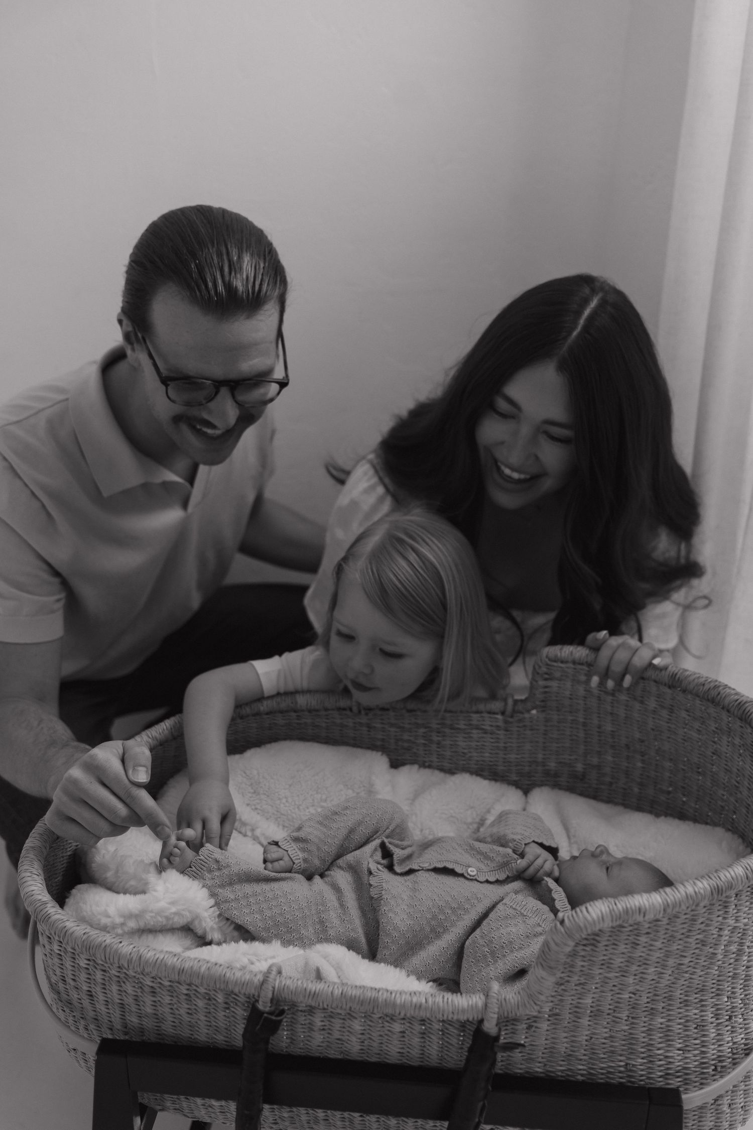 Black and white photo of family with toddler placing toy in newborn's wicker bassinet.