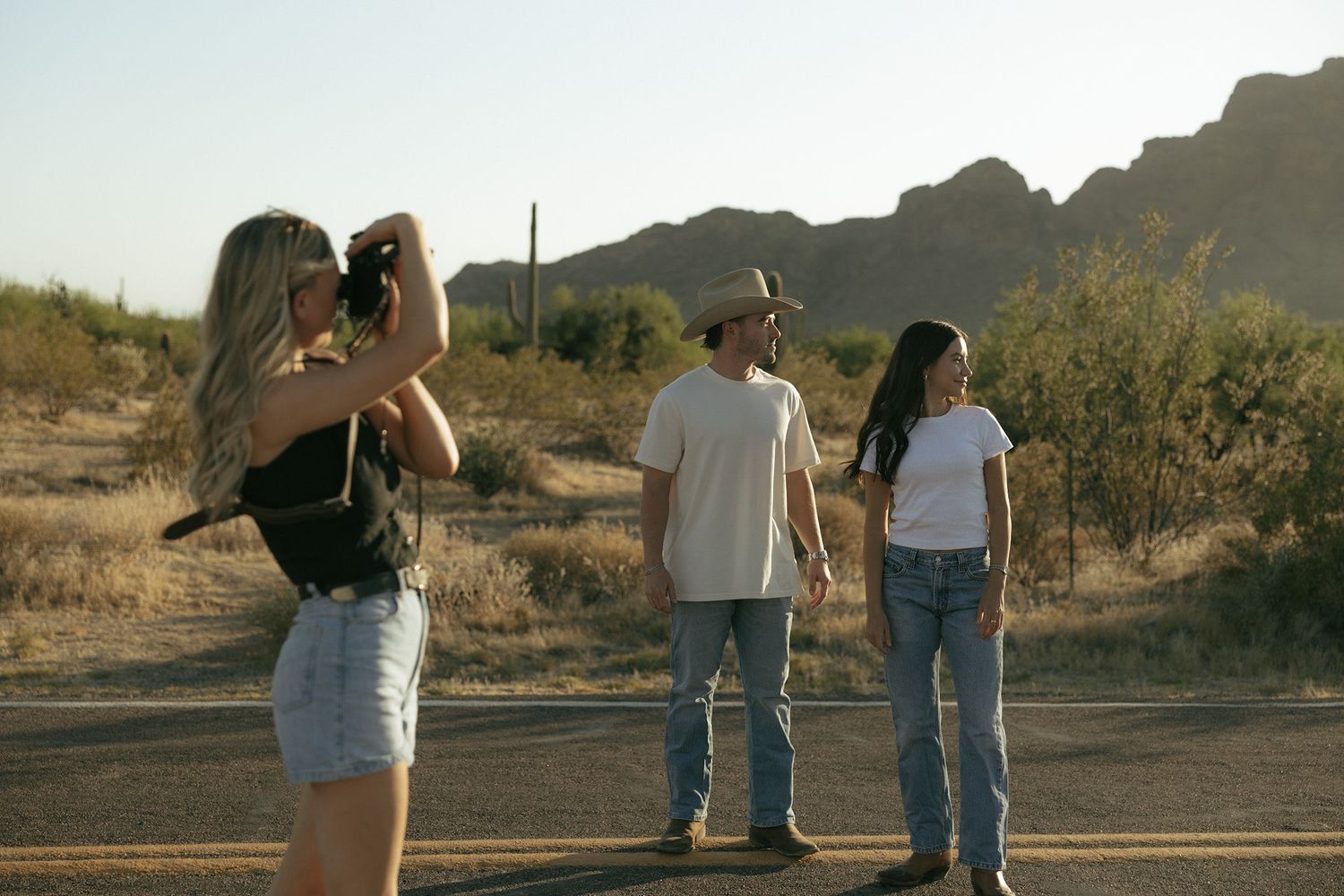Three people on a desert road, one woman photographing two others with mountains in the background.
