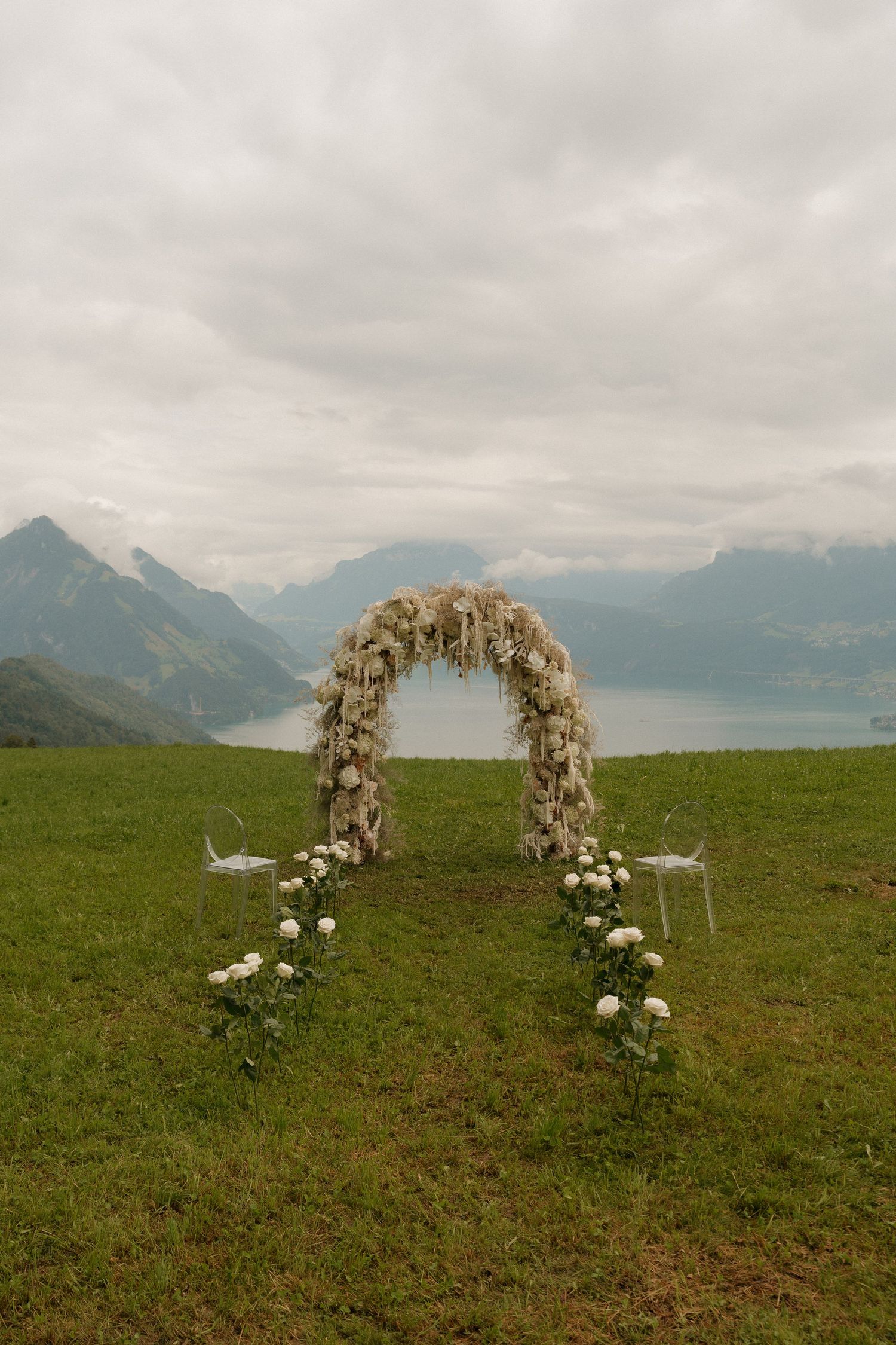 Floral wedding arch with white blooms on green hillside overlooking misty Alpine lake and mountains.
