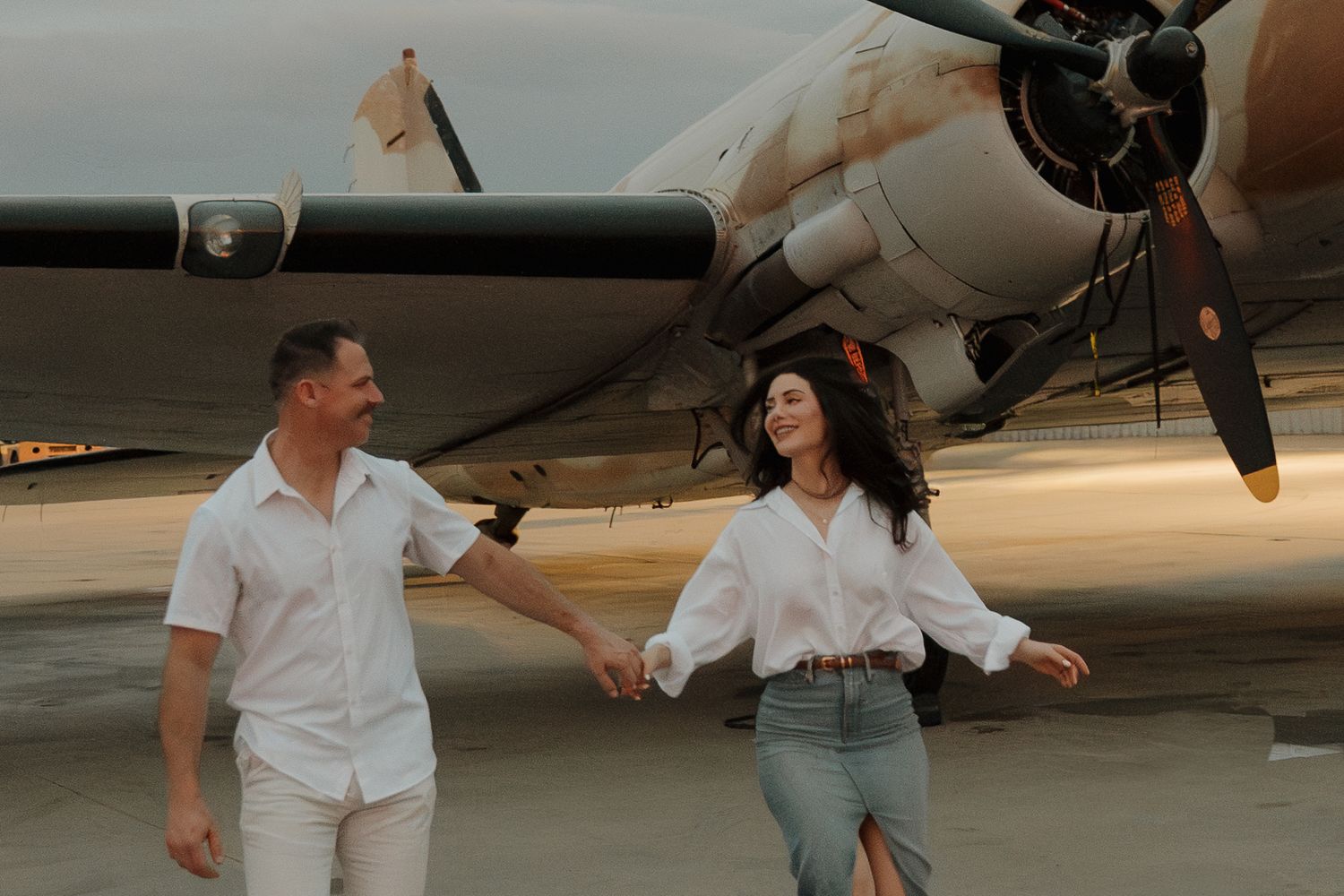 Couple holding hands and laughing in front of a vintage propeller airplane on a runway at golden hour.