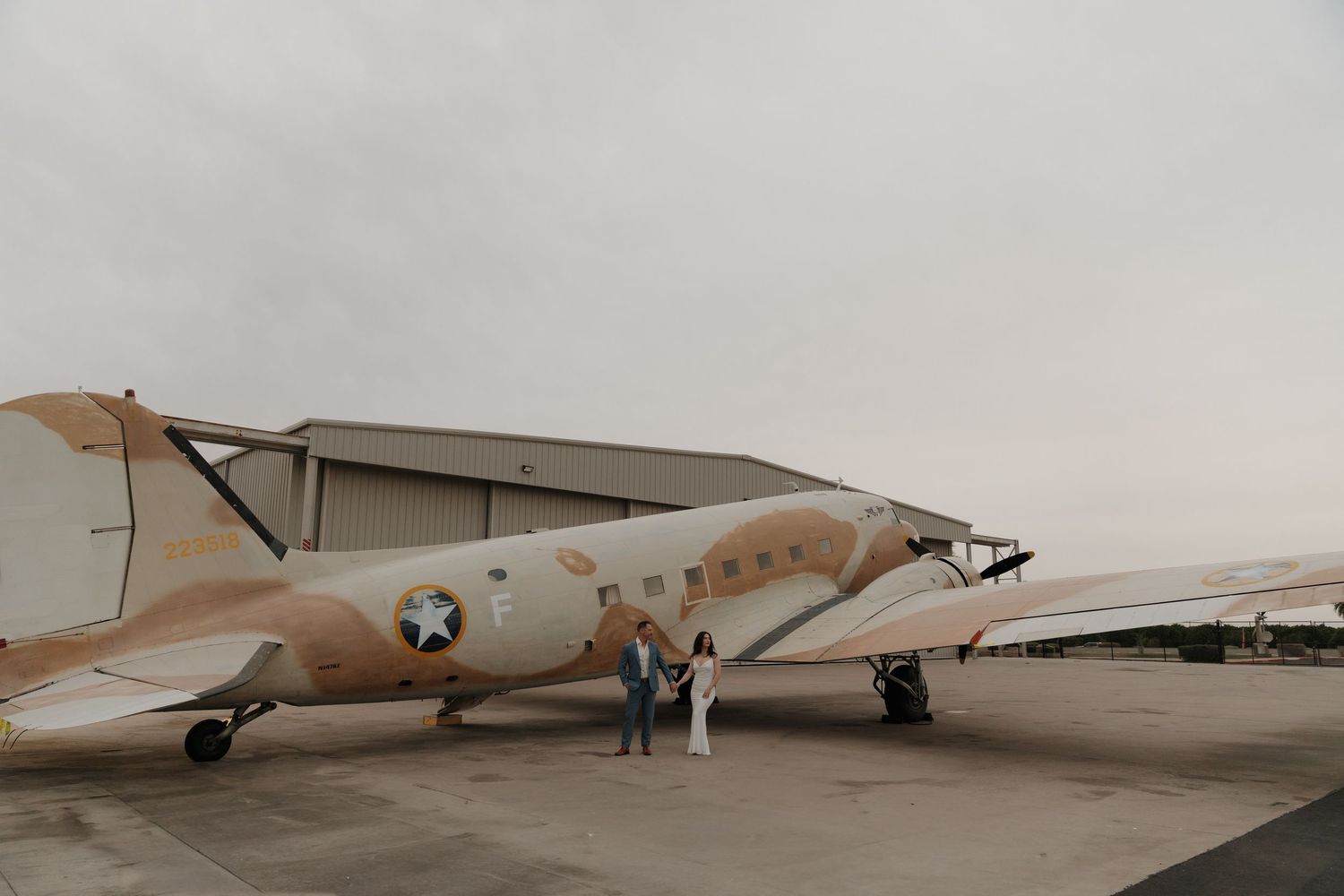 Couple poses beside a vintage WWII-era twin-engine aircraft on a tarmac with a hangar in the background.