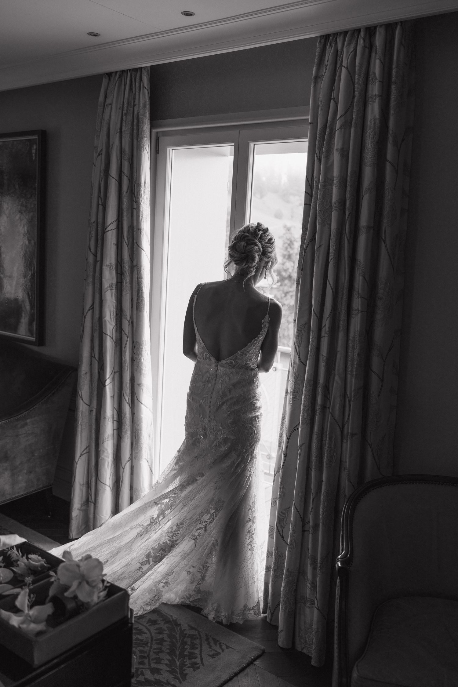 Black and white silhouette of bride in backless lace wedding gown standing before large hotel window.