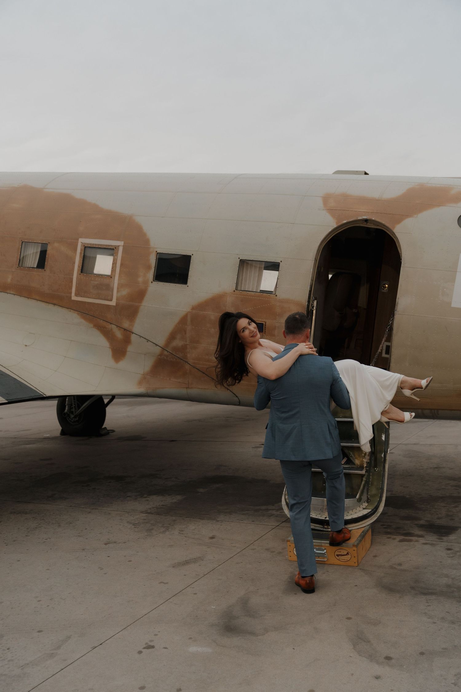 Groom carries bride toward vintage airplane door in romantic outdoor wedding photo shoot on tarmac.