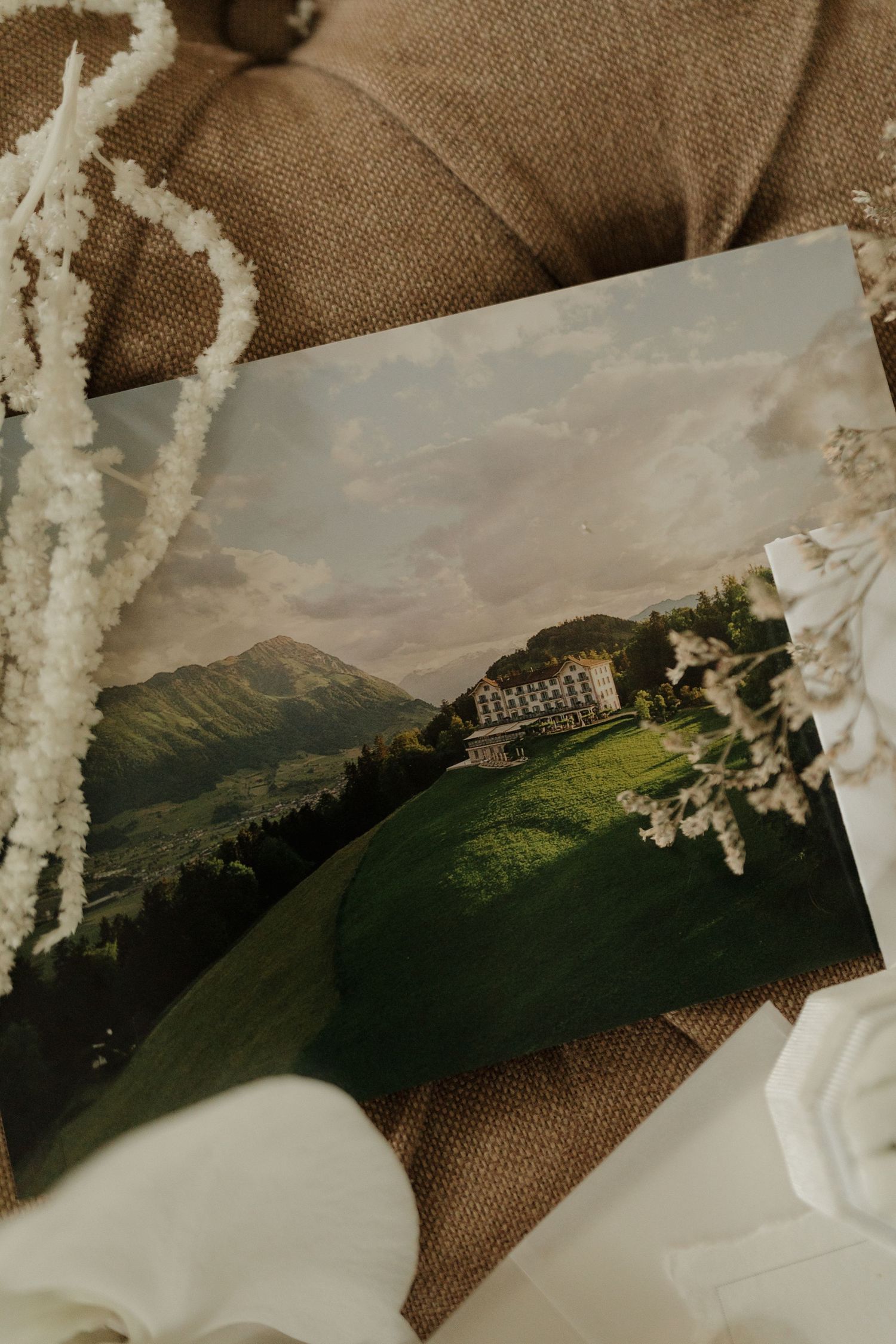 Vintage postcard of grand Alpine hotel on green hillside with mountain backdrop and white florals.