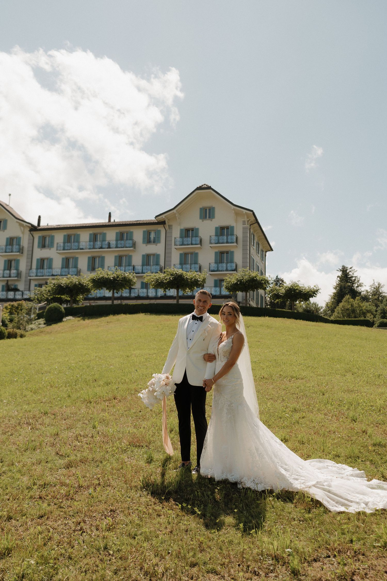 Bride and groom posing on green lawn before elegant hotel on sunny wedding day.