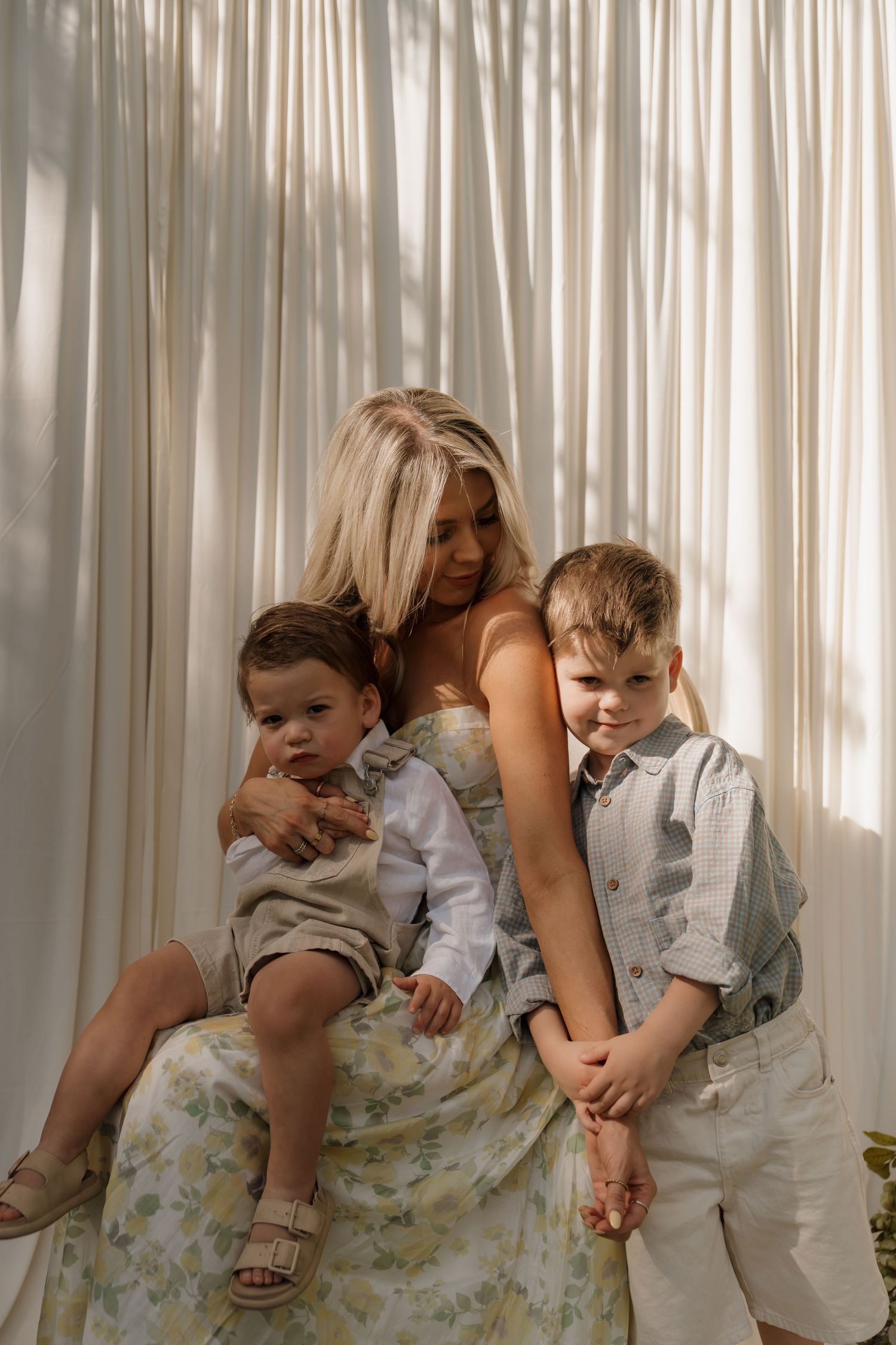 Blonde mother in floral dress sits with two young sons against sheer white curtain backdrop.