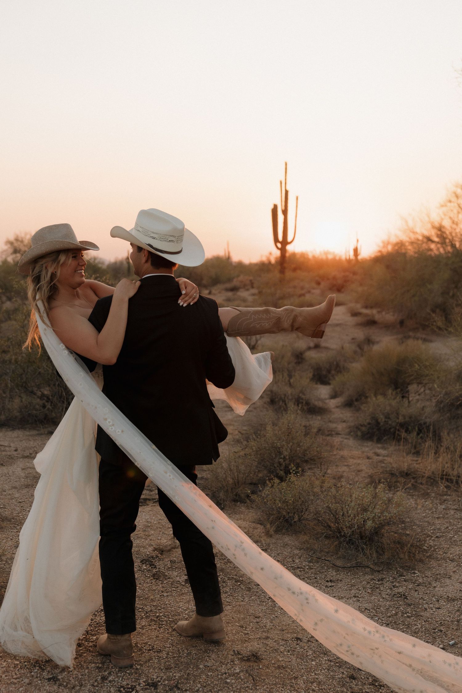 Groom carrying bride in cowboy hats at desert sunset with saguaro cactus in background.