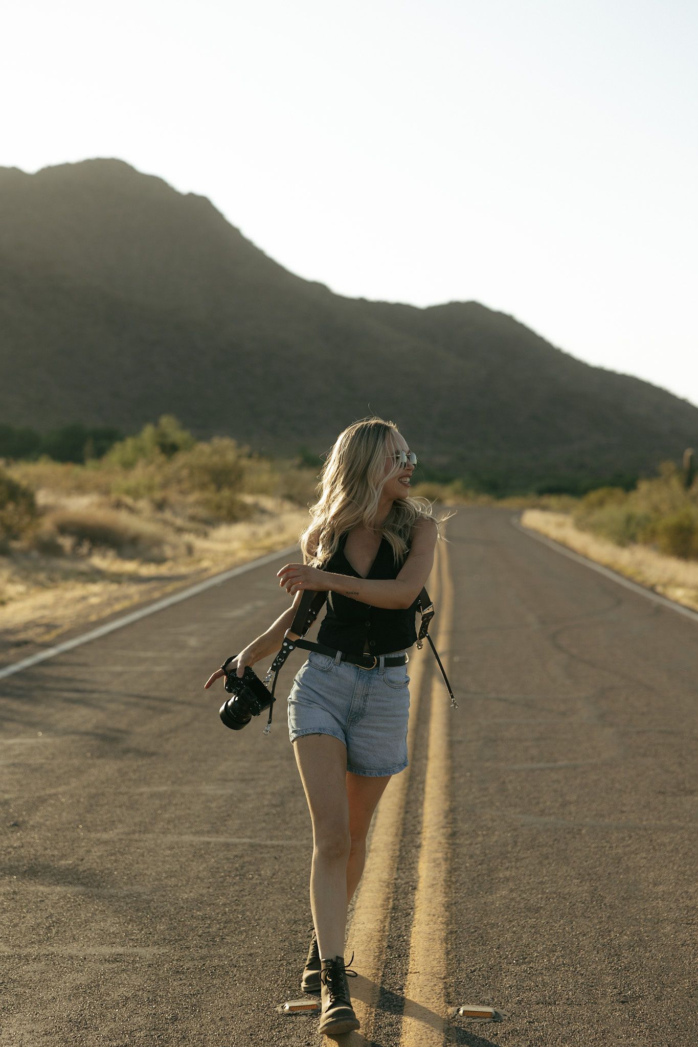 Blonde woman in denim shorts holding camera gear standing on desert road with mountain backdrop in golden hour light.