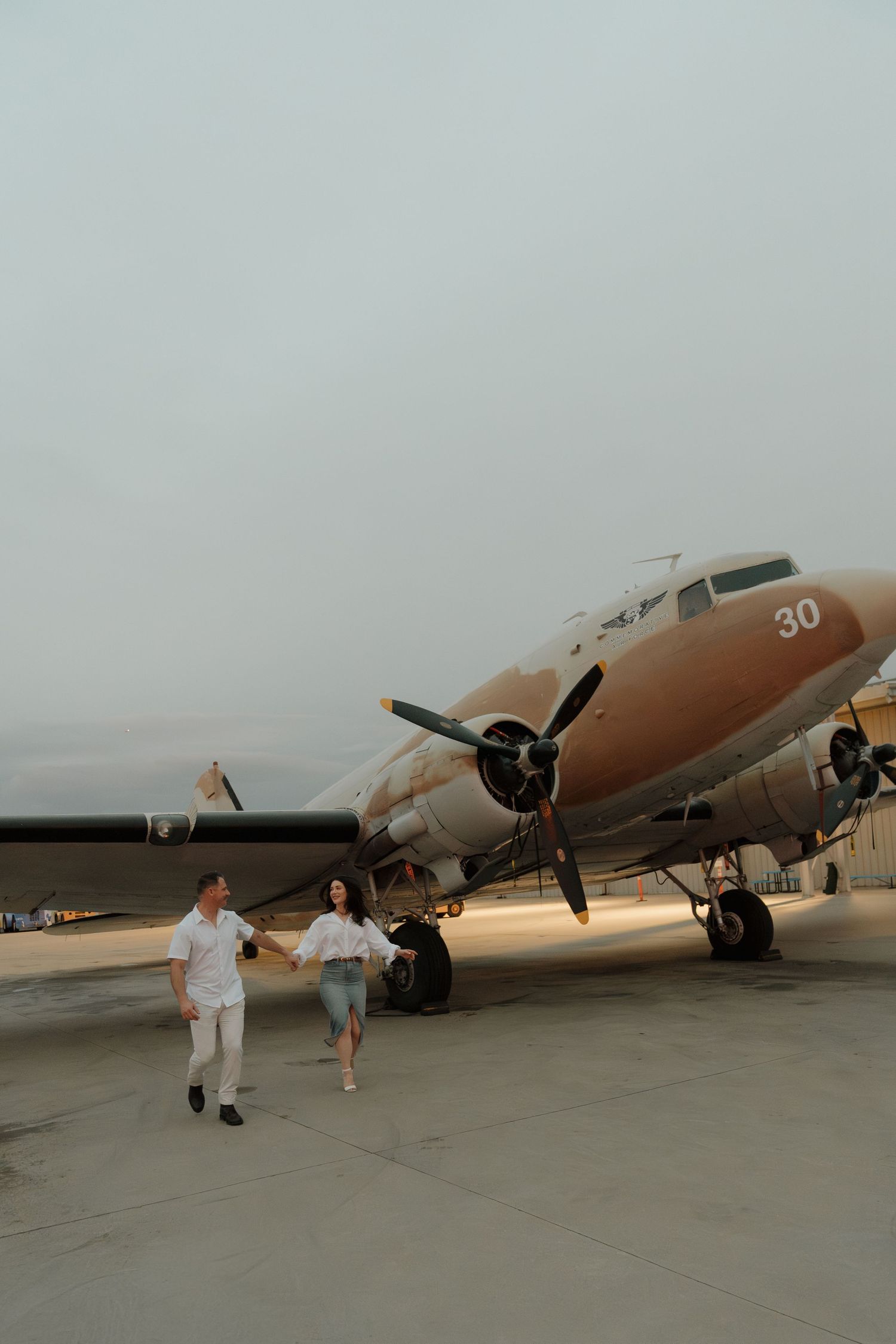 Couple walking hand in hand on tarmac beside vintage propeller aircraft numbered 30 at dusk.