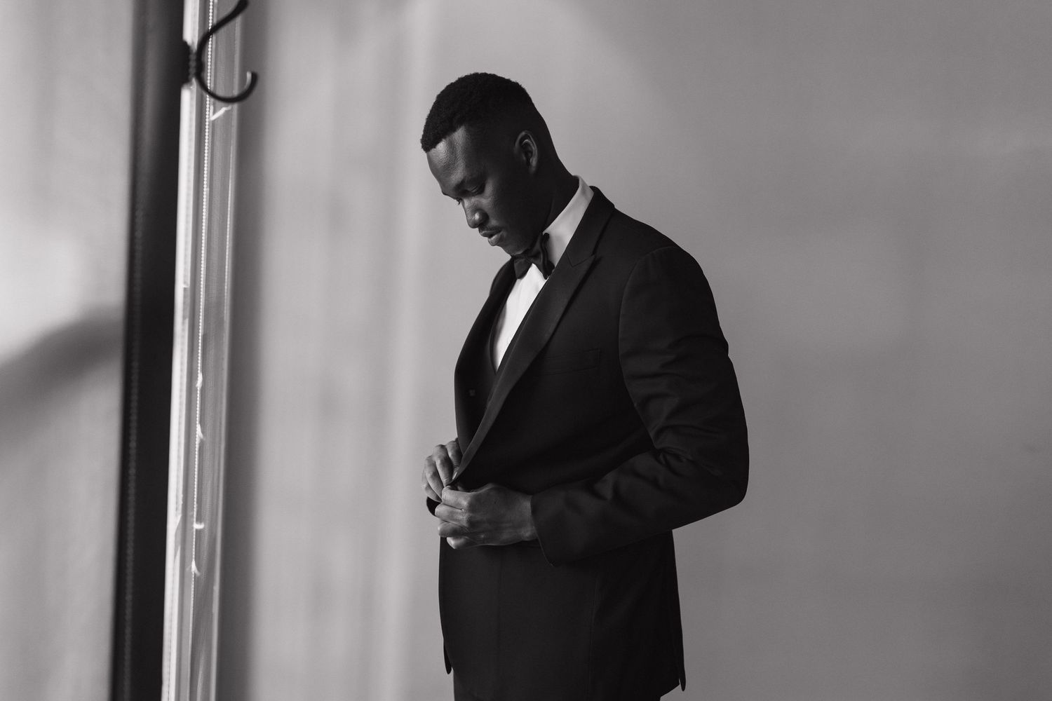 Black and white portrait of man in tuxedo buttoning jacket by window.