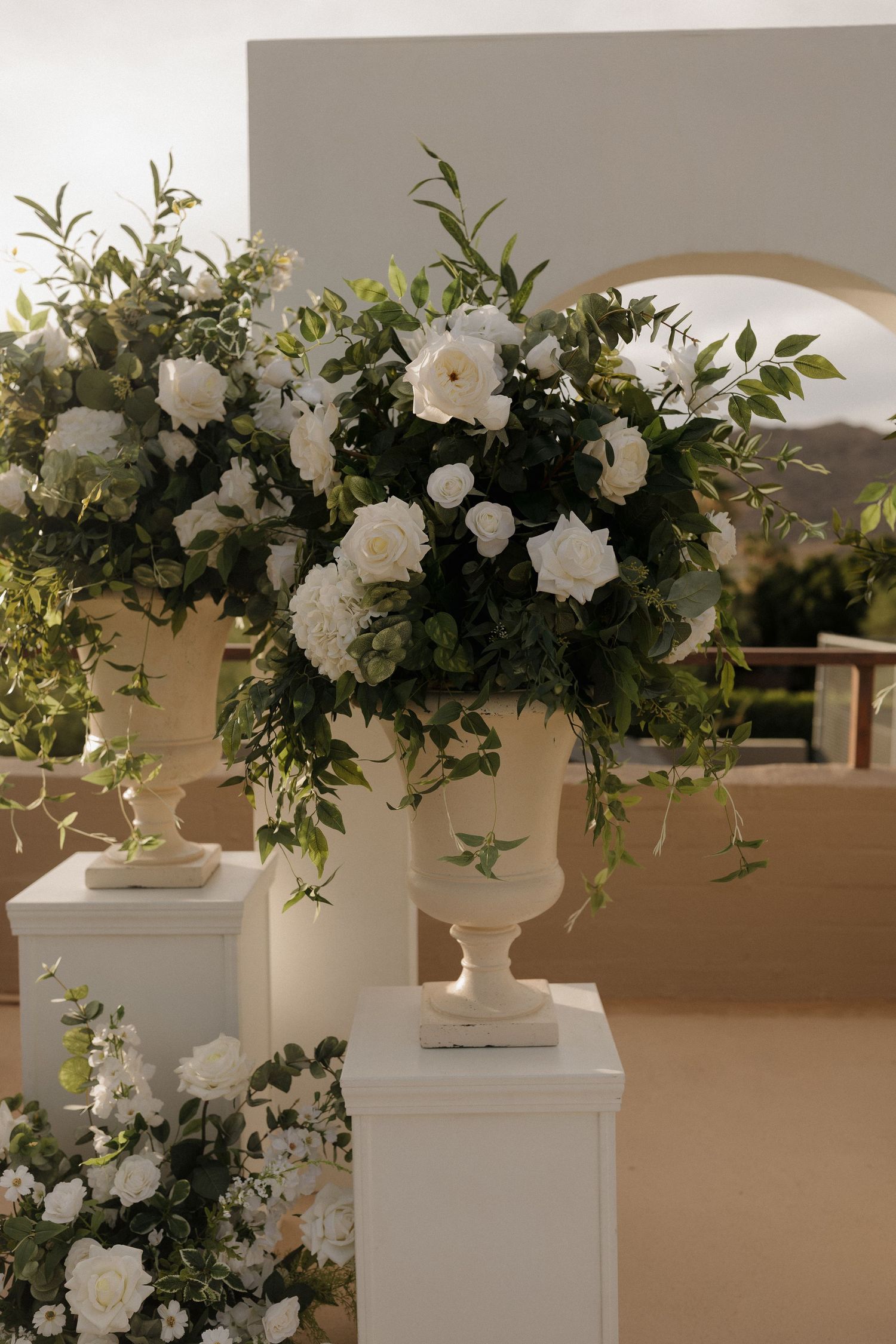 Elegant white floral arrangement with roses and greenery in a classic urn vase on a white pedestal.