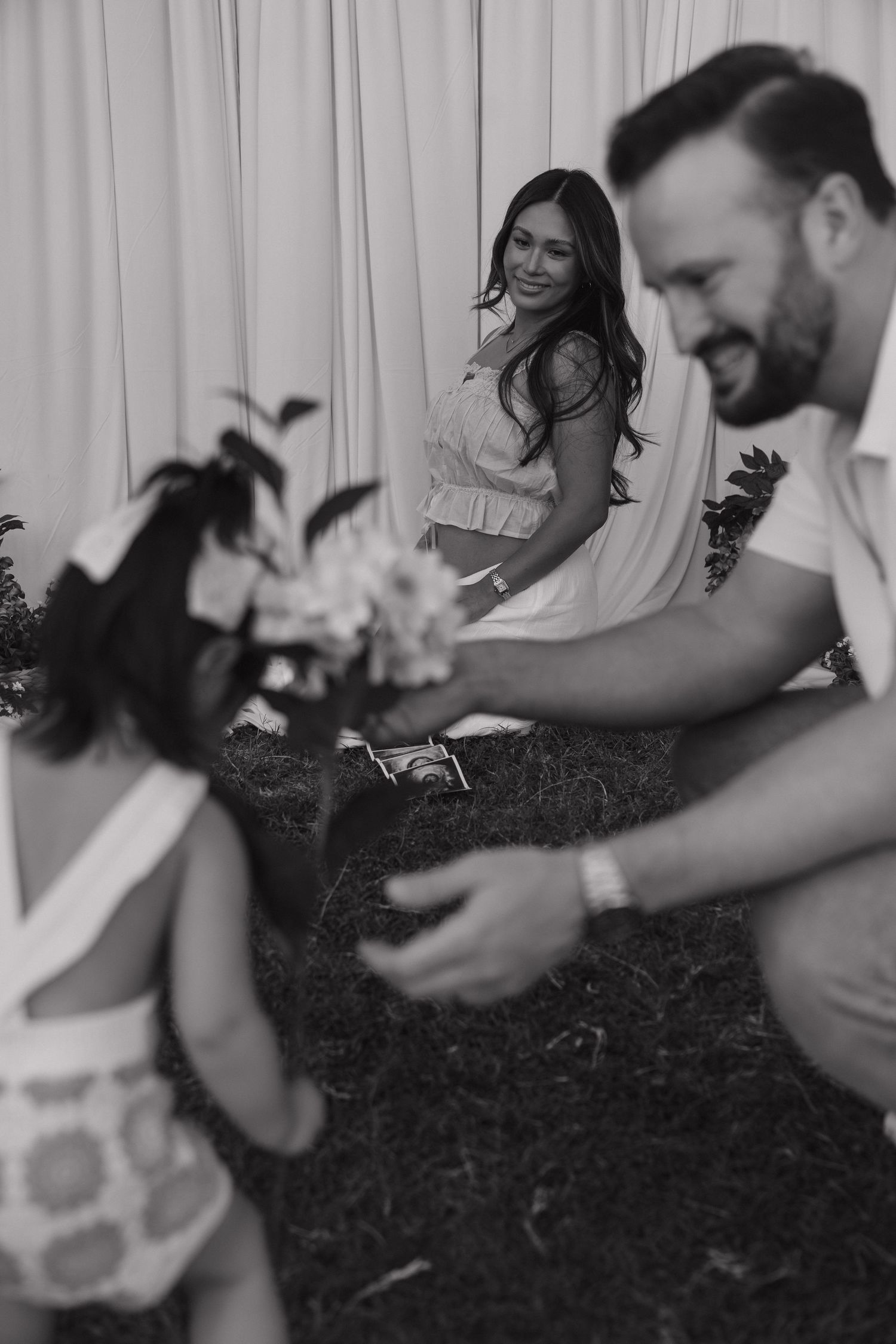Black and white photo of couple interacting with young flower girl at outdoor wedding ceremony.