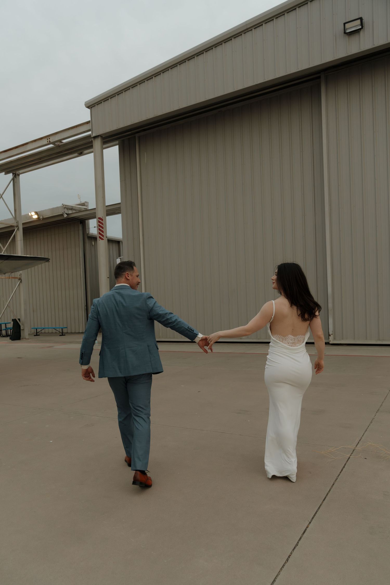 Bride and groom walking hand in hand toward an industrial hangar venue, she in a white backless gown, he in a blue suit.