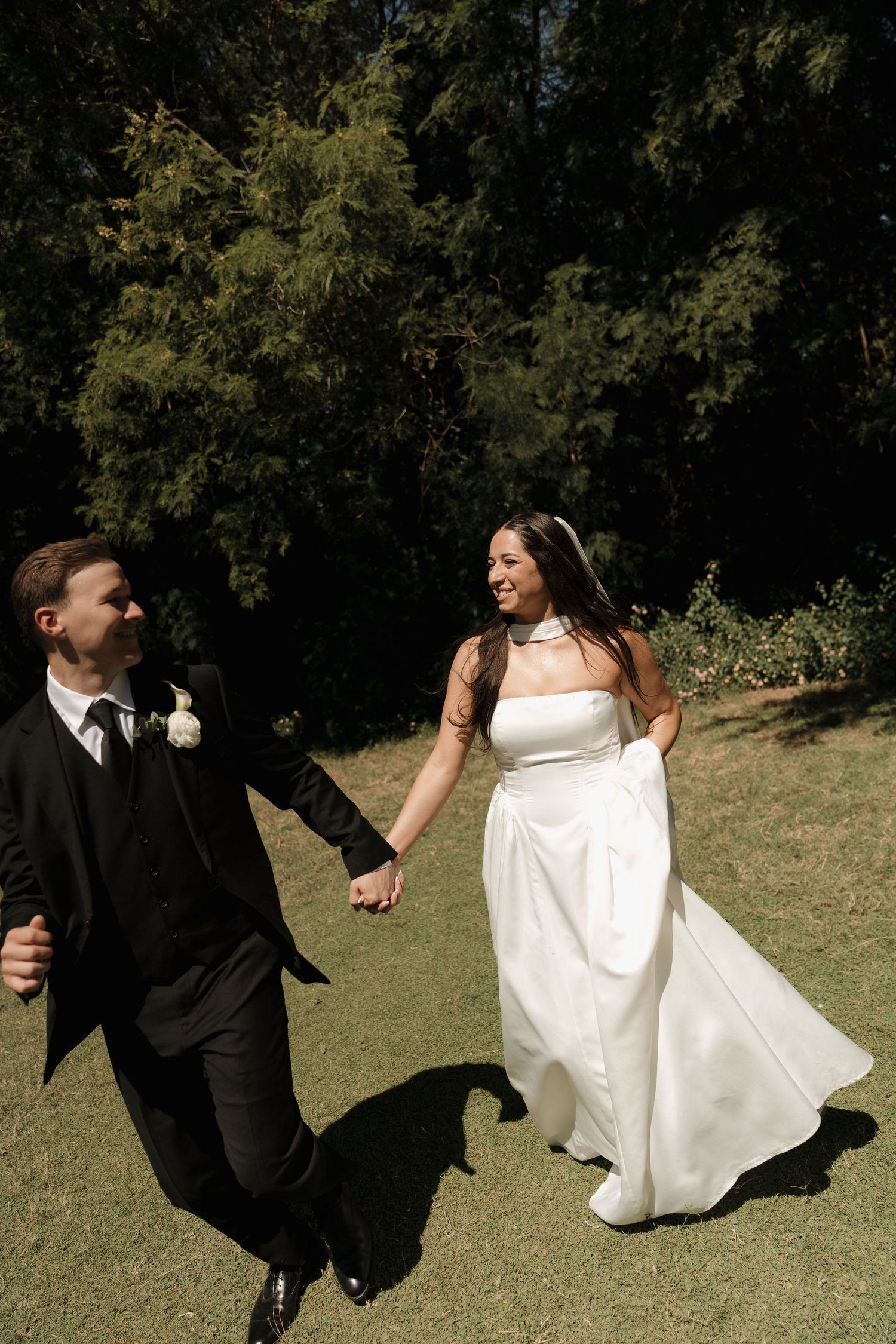 Bride and groom holding hands walking on grass, bride in white gown, groom in black suit.