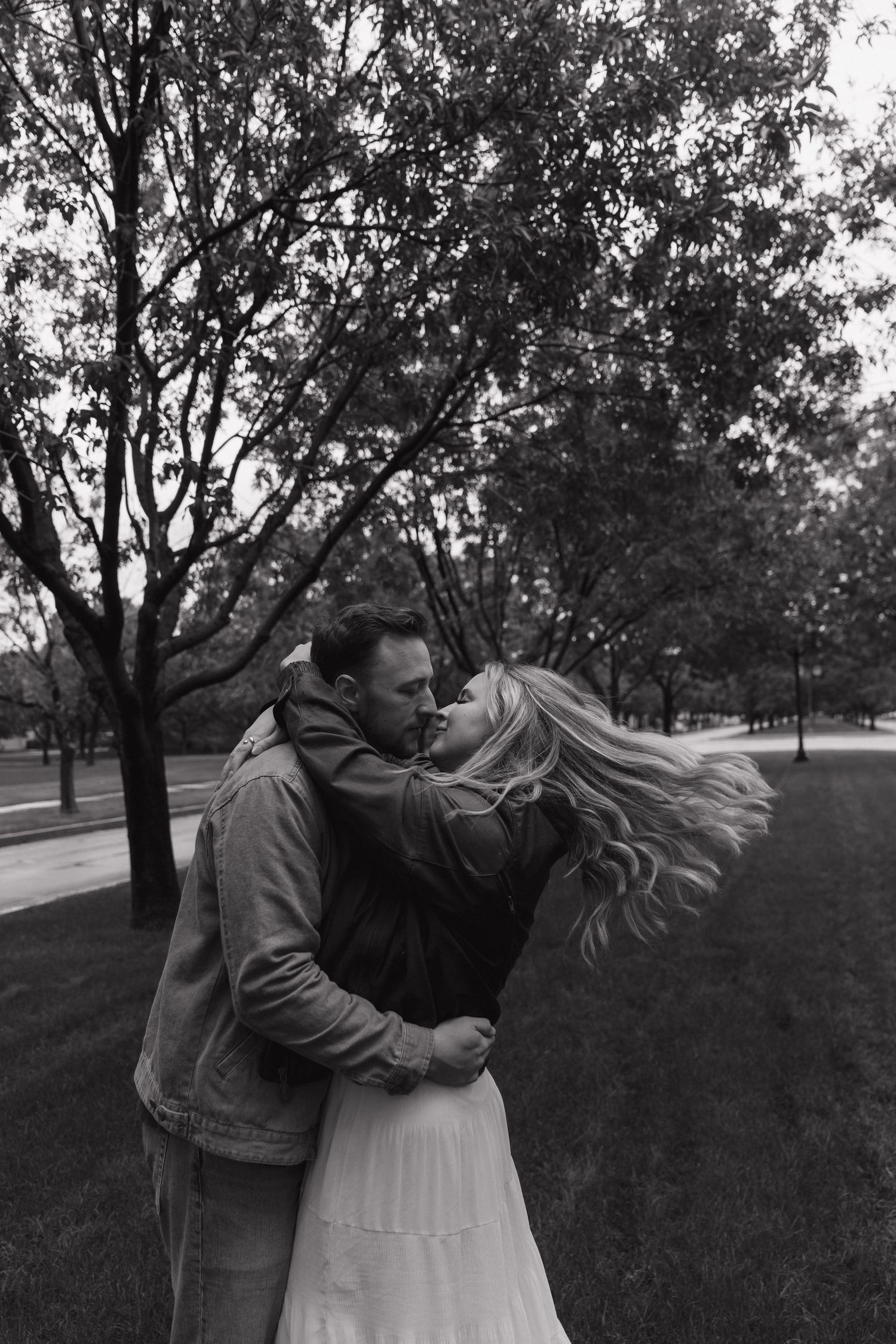 Black and white photo of a couple kissing in a park, woman's blonde hair blowing in the wind.