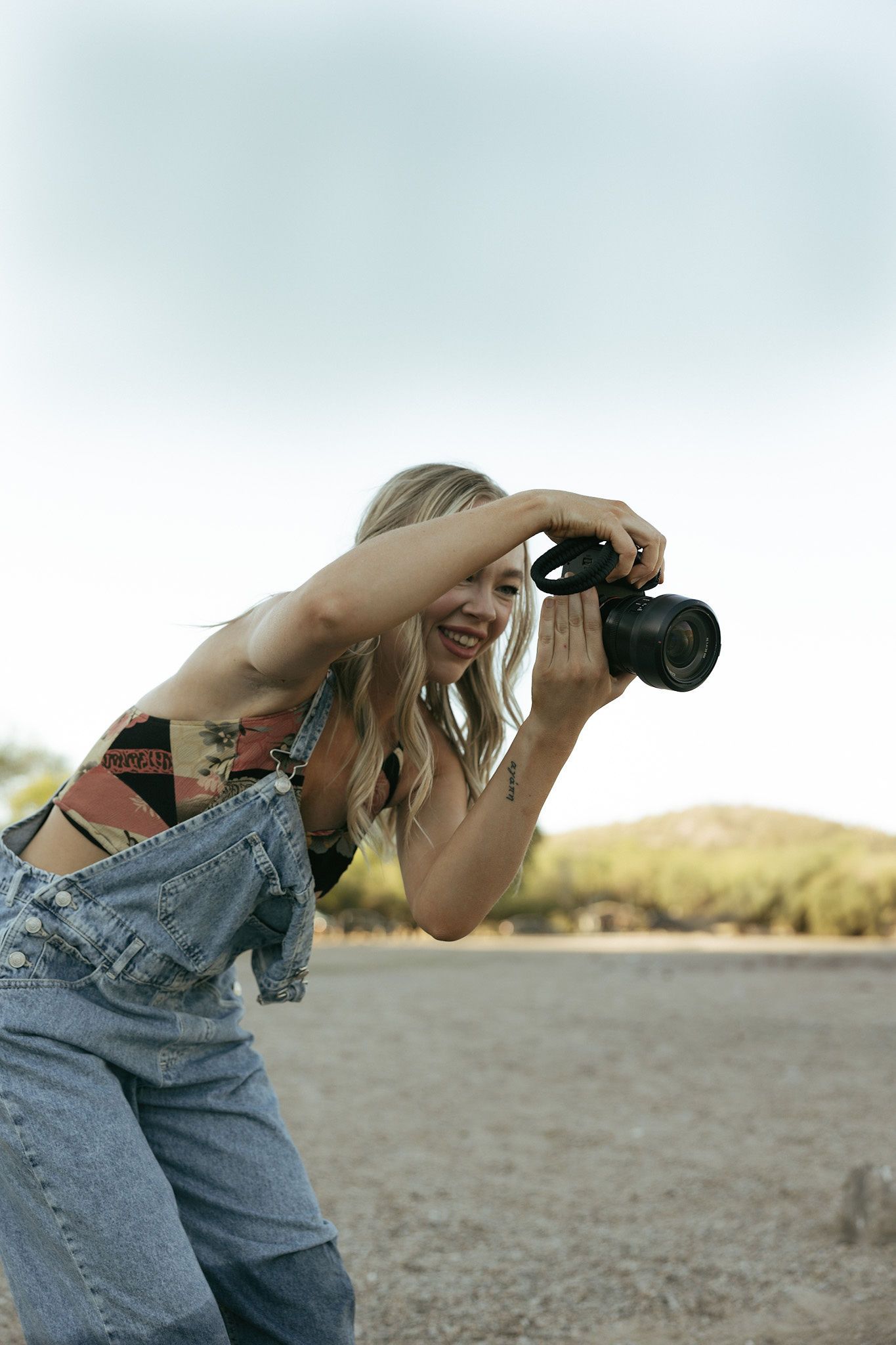 Smiling blonde woman in denim overalls crouching outdoors while taking photos with a DSLR camera in a desert landscape.