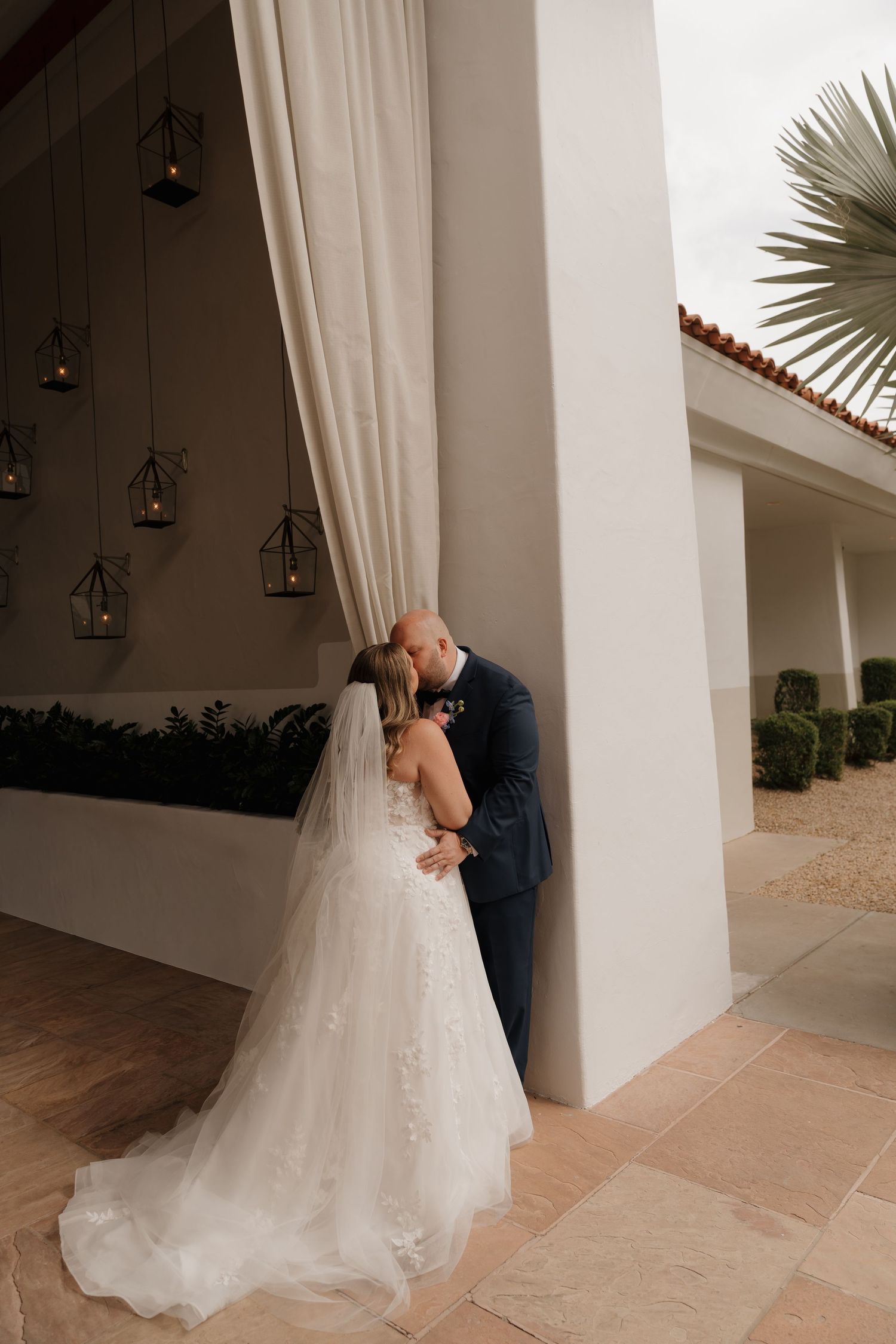 Bride and groom sharing a kiss beside white column with hanging lanterns and veil trailing.