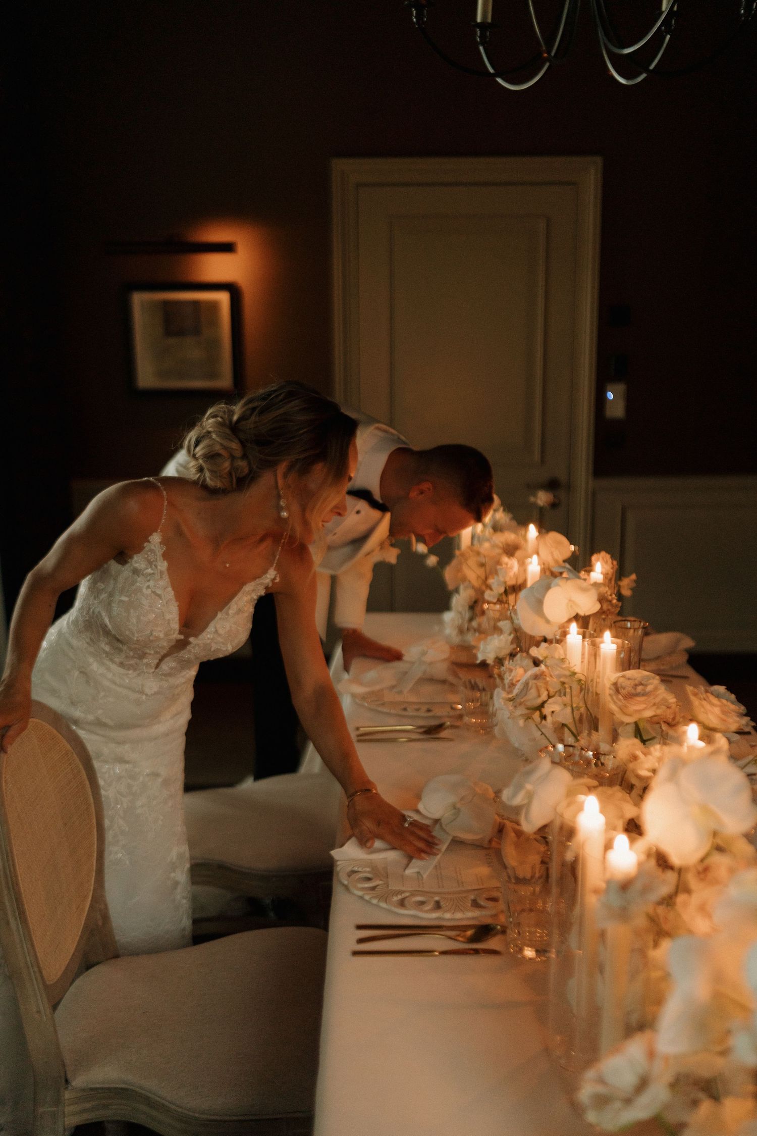 Bride and groom lean over candlelit wedding reception table adorned with white flowers in dim room.