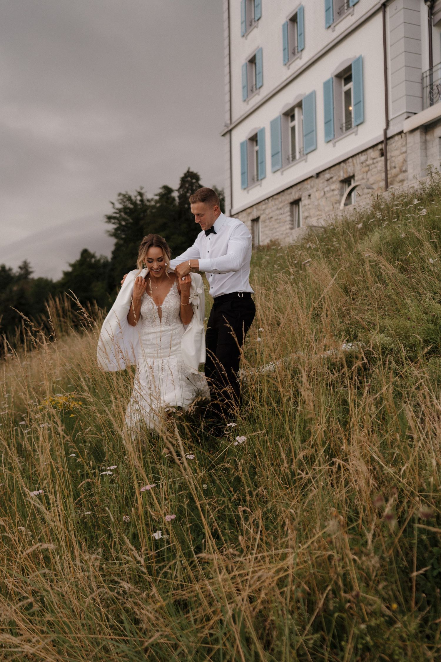 Groom helping bride with her veil while walking through tall wildflower meadow near elegant villa.