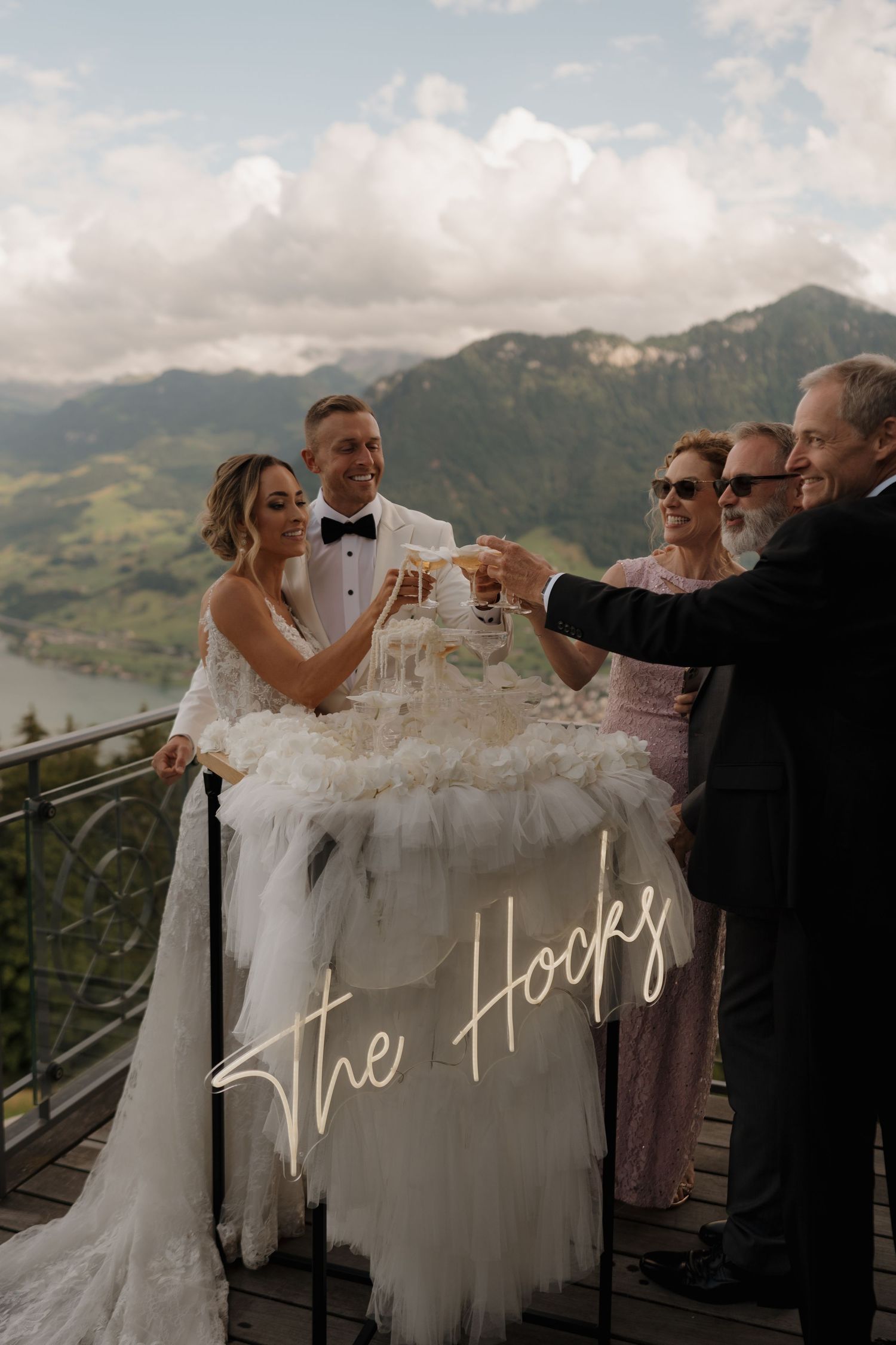 Bride and groom celebrating with guests, raising champagne glasses on a balcony with alpine lake views.