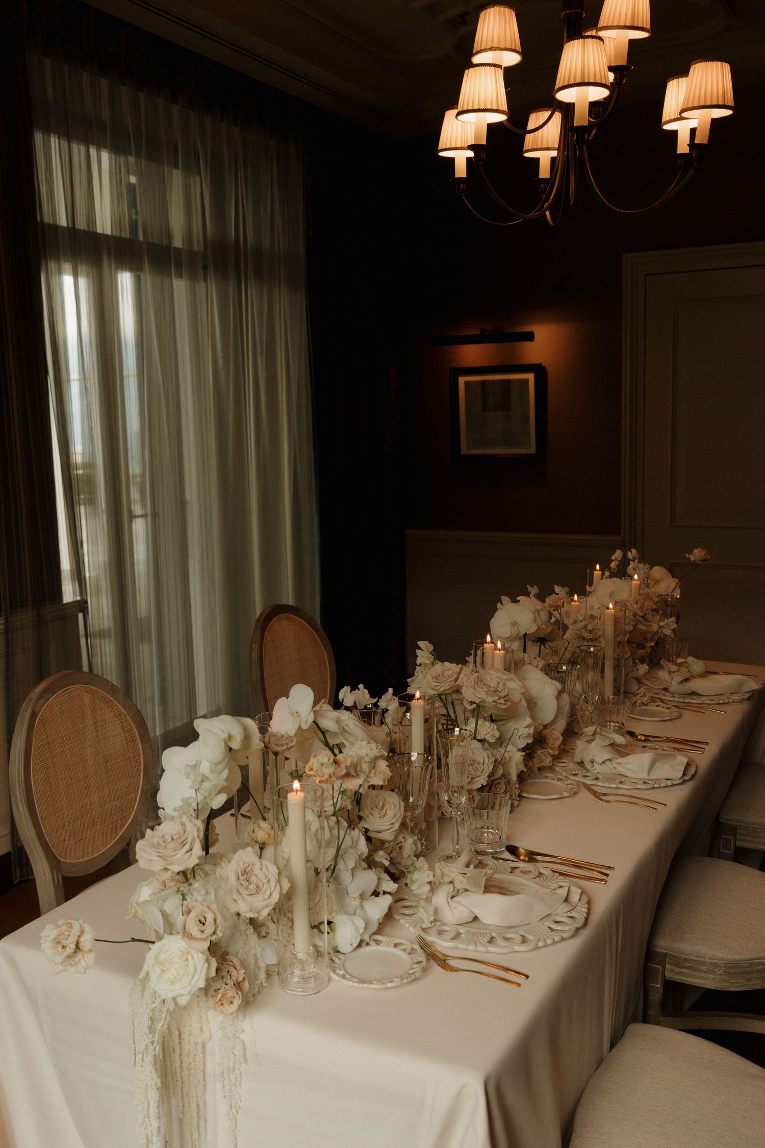 Luxurious wedding reception table with white orchids, roses, candles, and gold accents in dark room.