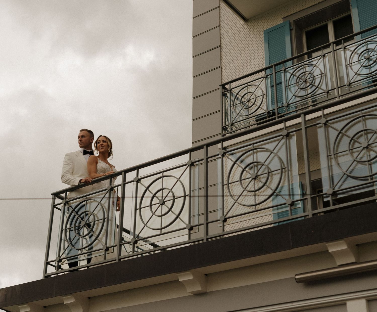 Wedding couple standing together on ornate balcony of elegant building under cloudy sky.