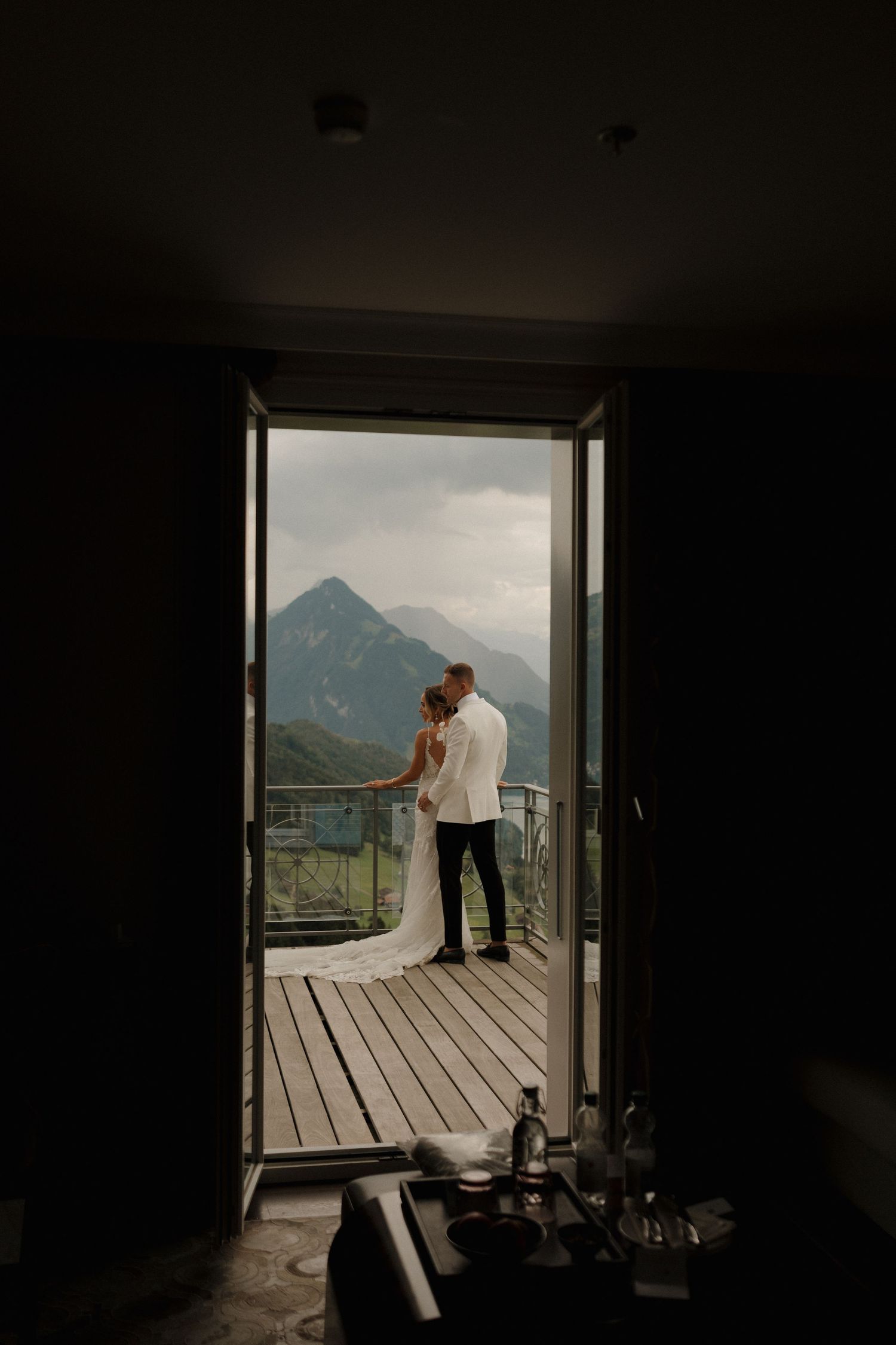 Romantic silhouette of bride and groom kissing on balcony framed by dark doorway with mountain views.