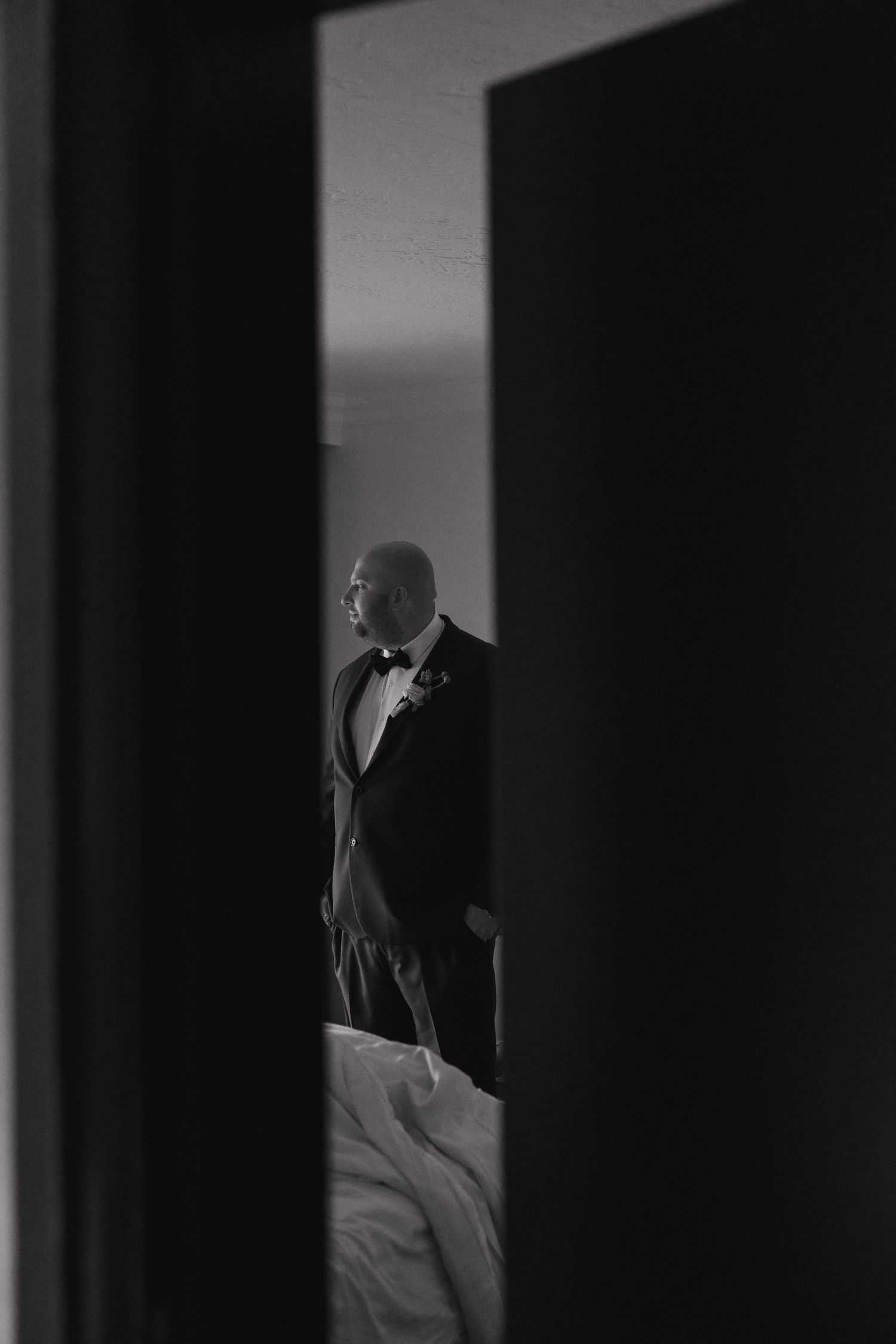 Dramatic black and white photo of groom in tuxedo framed by dark doorway in moody lighting.