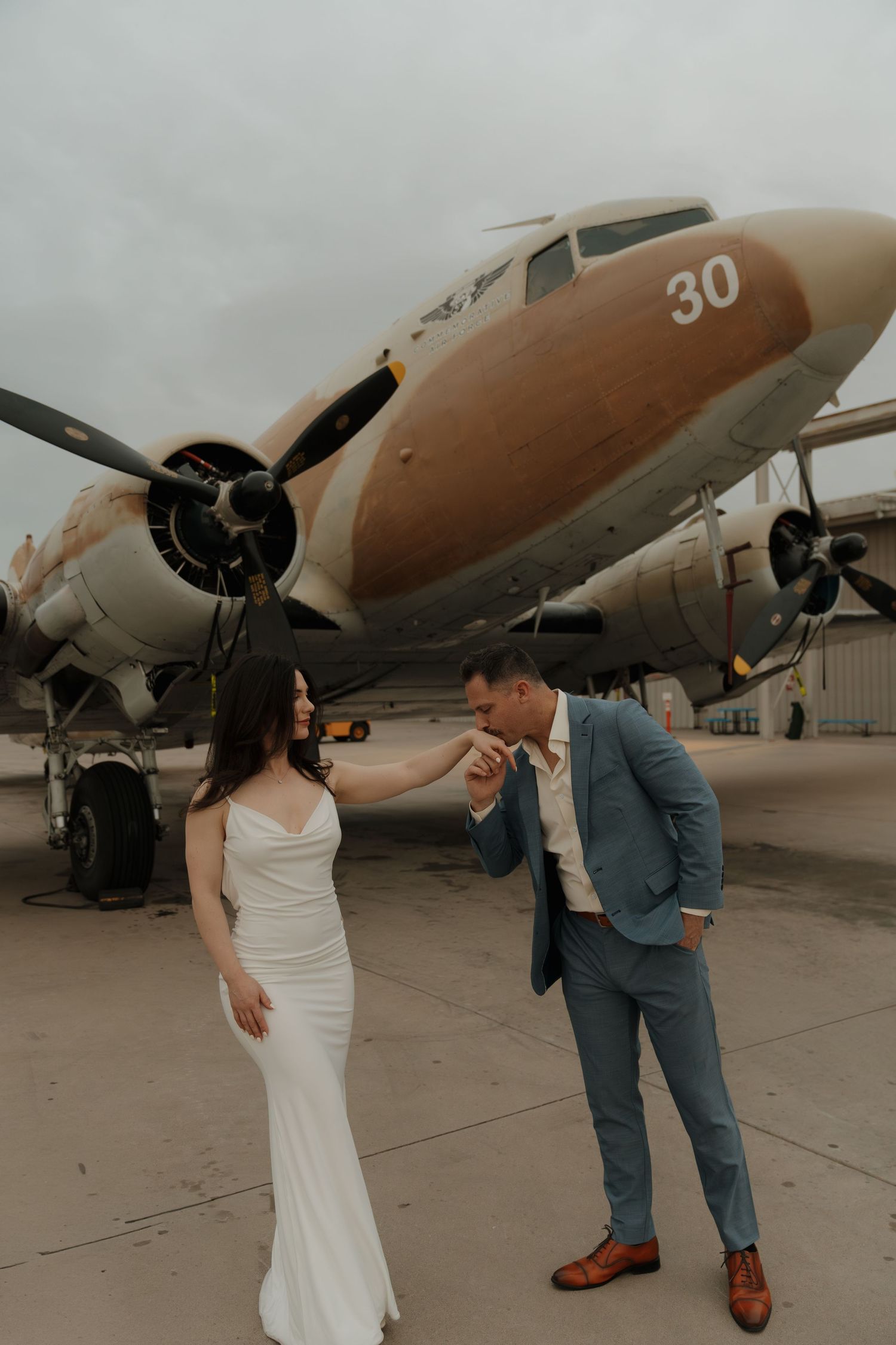 Groom kissing bride's hand in front of a rustic vintage DC-3 airplane numbered 30 on a tarmac.