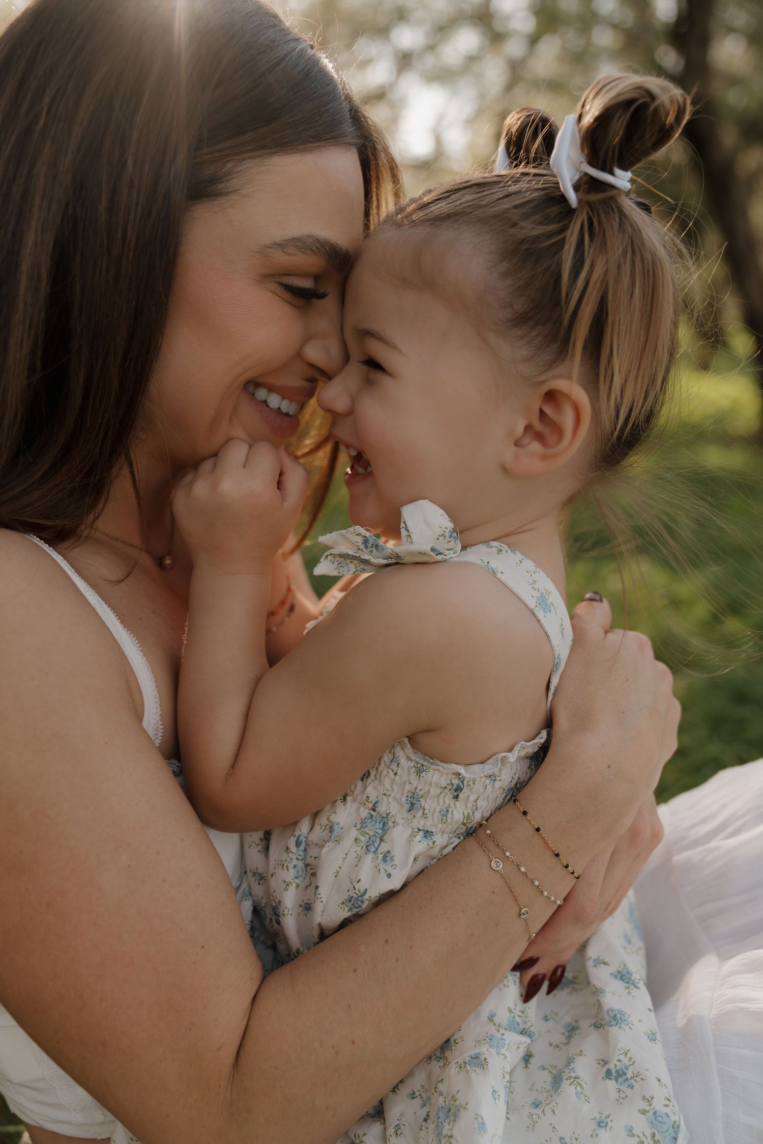 Joyful mother laughing and holding toddler girl in floral dress outdoors in sunlight.