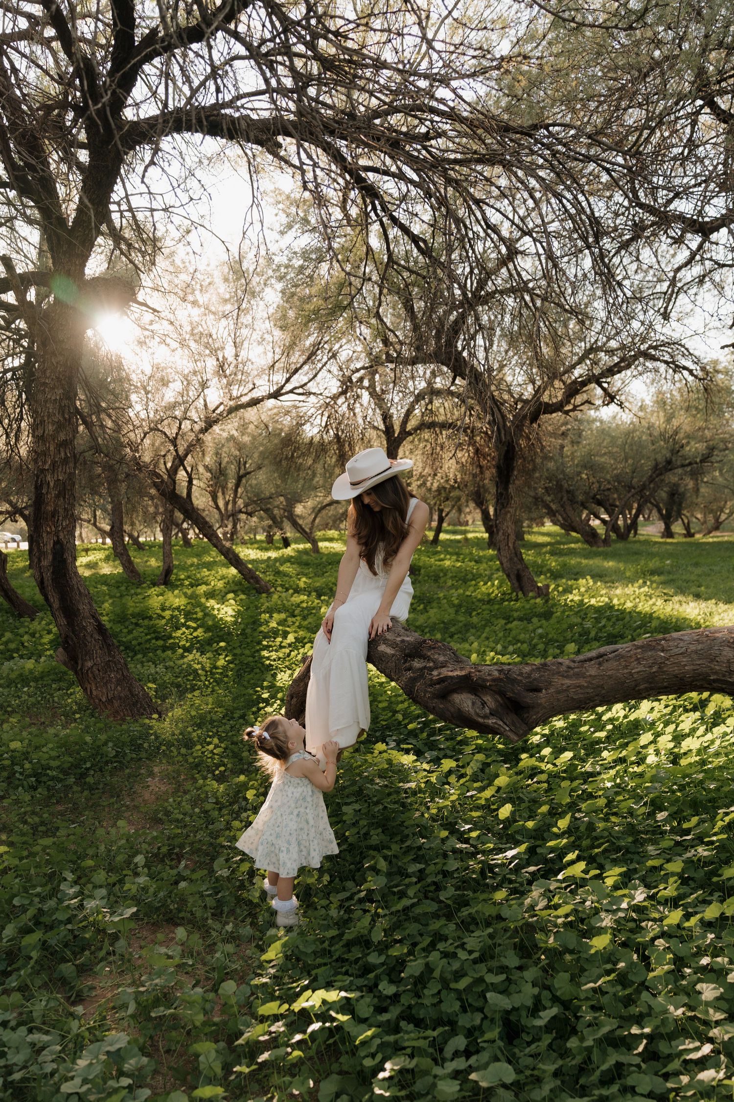 Mother in white dress and sun hat holding daughter's hand in sunlit green park.