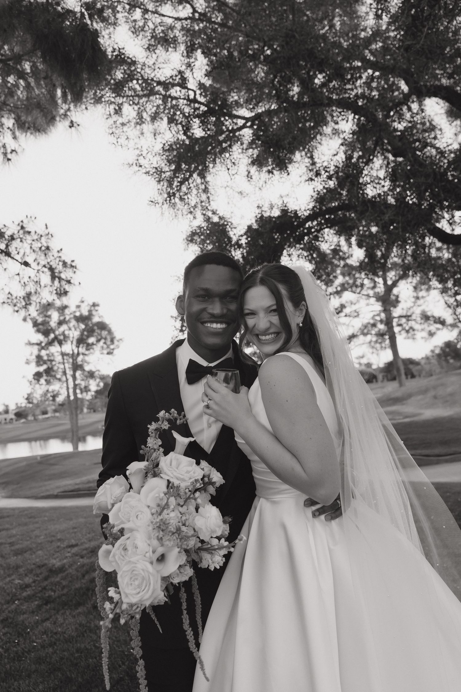 Black and white portrait of smiling newlywed couple holding bouquet under trees near a pond.