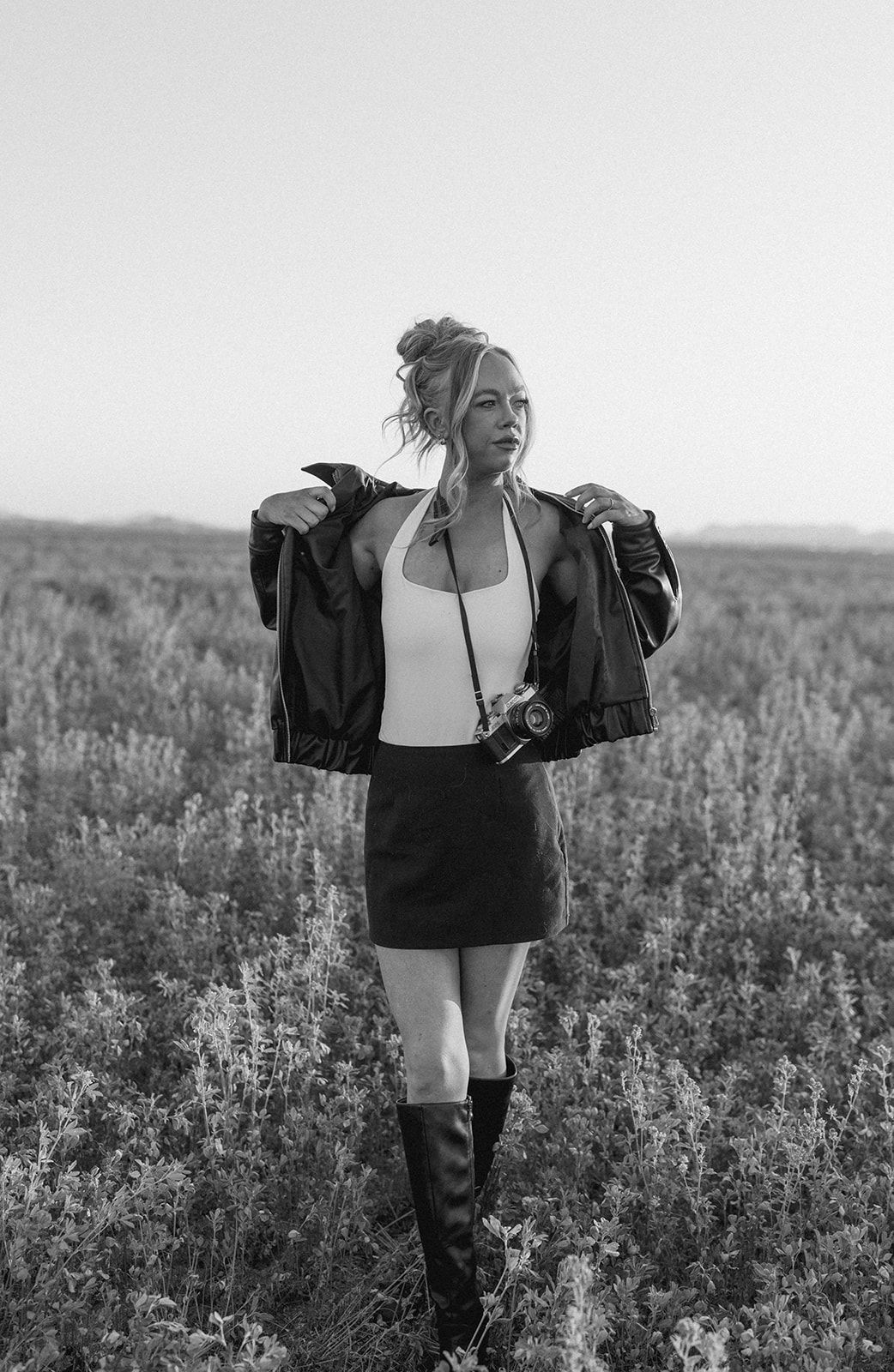 Black and white portrait of blonde woman in leather jacket holding vintage camera in open field.