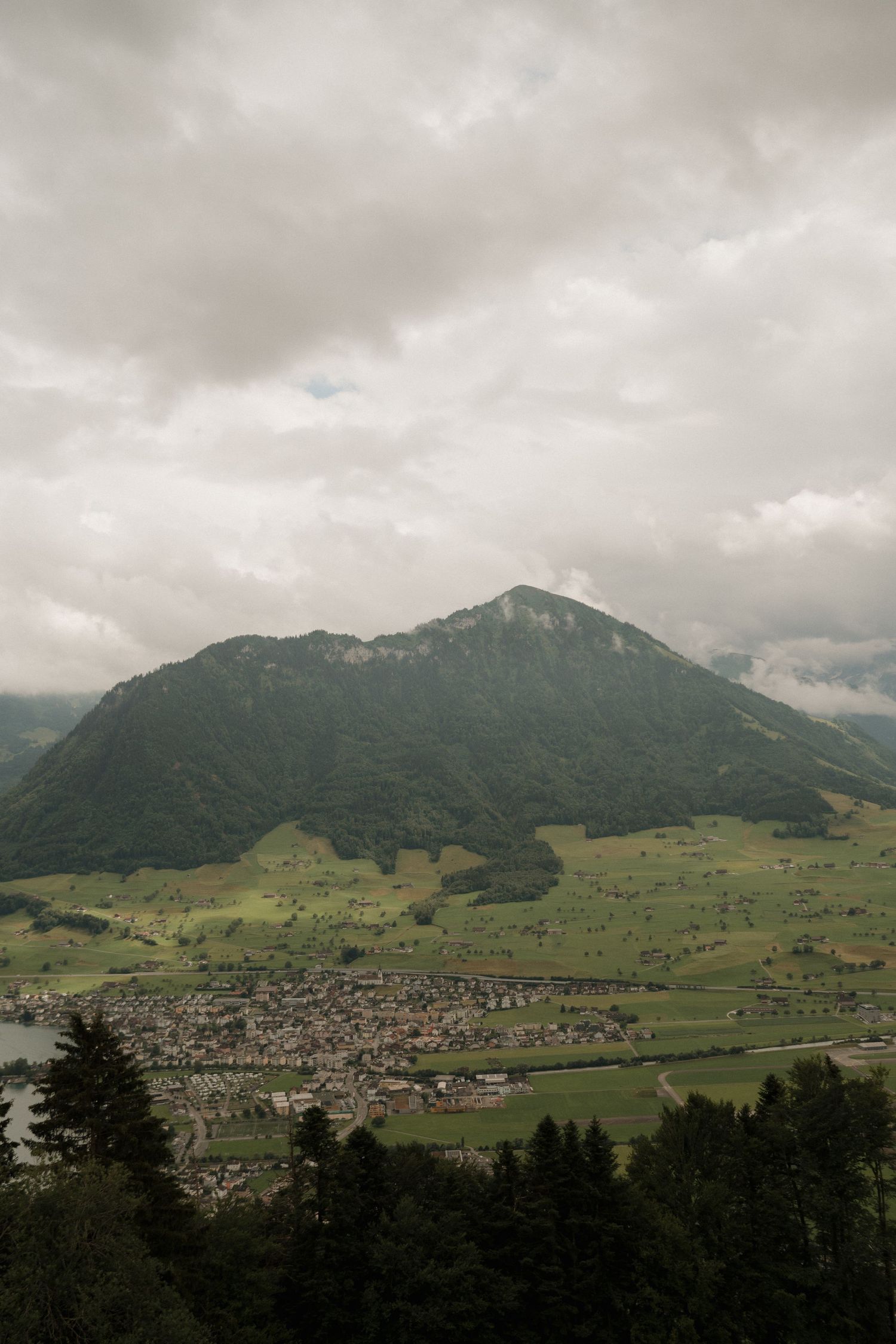 Dramatic aerial view of Alpine valley town nestled below large green mountain under cloudy sky.